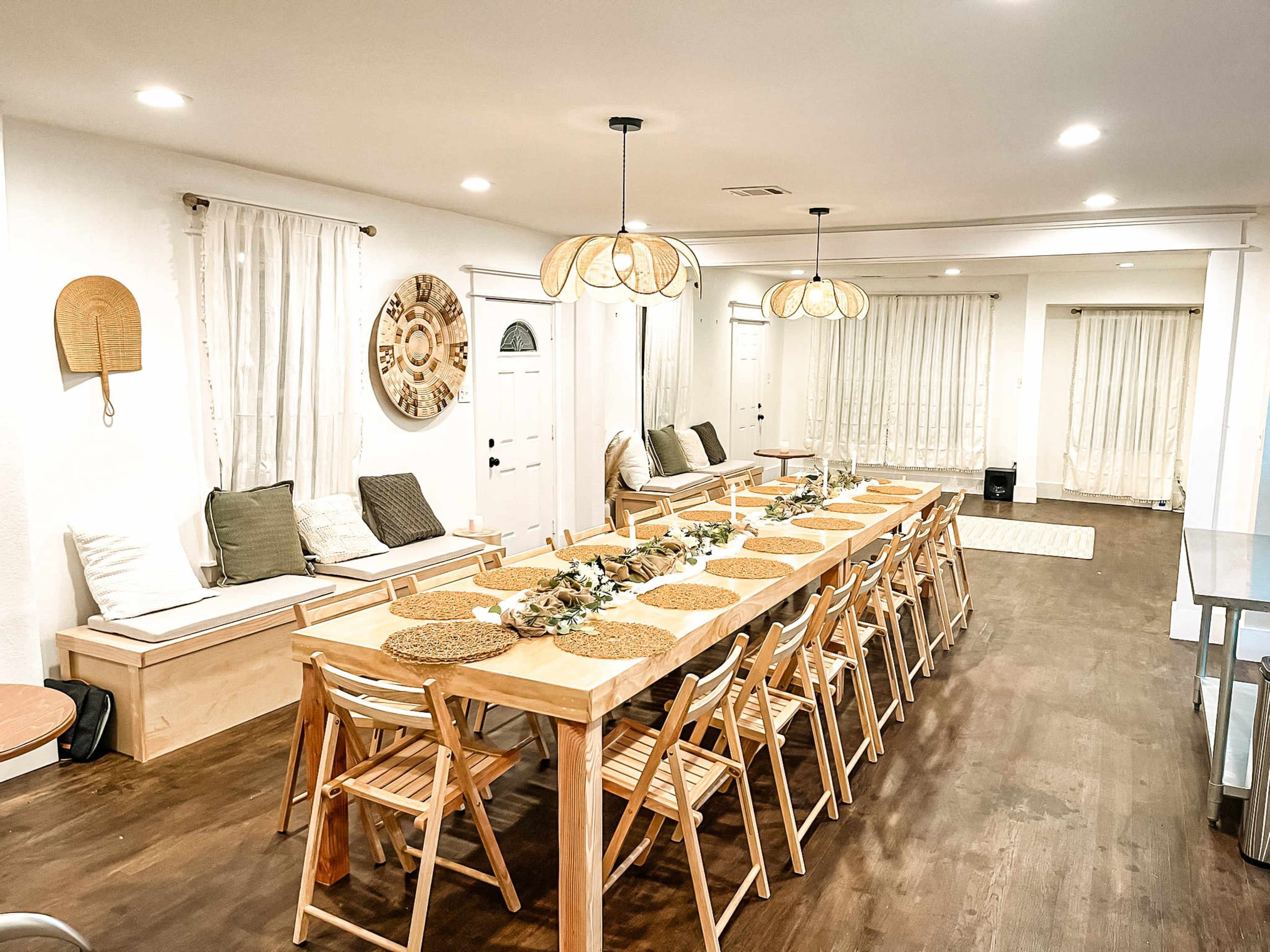 A long wooden dining table with woven placemats is set for a meal in a well-lit dining room featuring white walls, large windows with sheer curtains, and a cozy seating area.