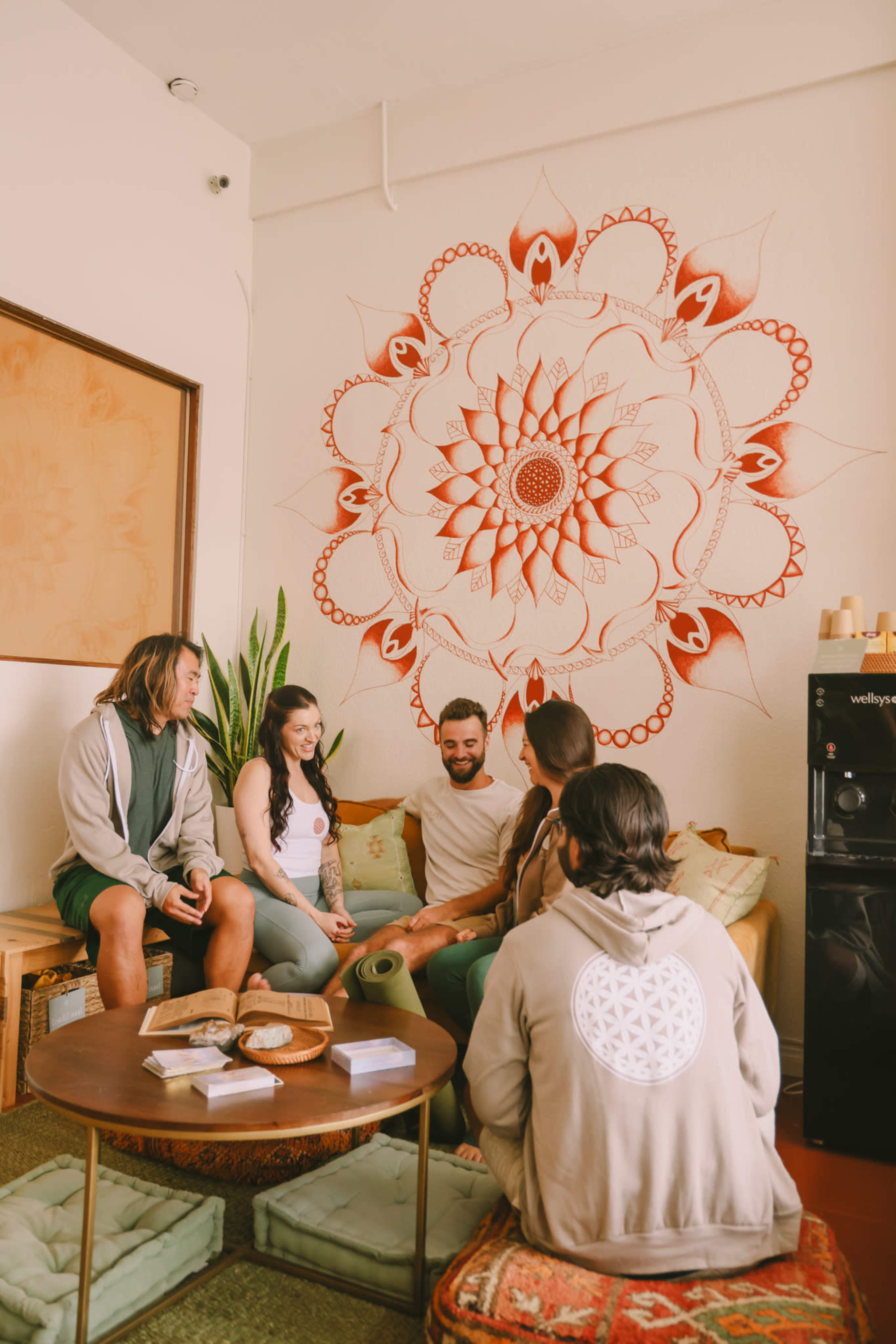 A group of five people is seated on cushions in a cozy room, with a large mandala mural on the wall behind them.