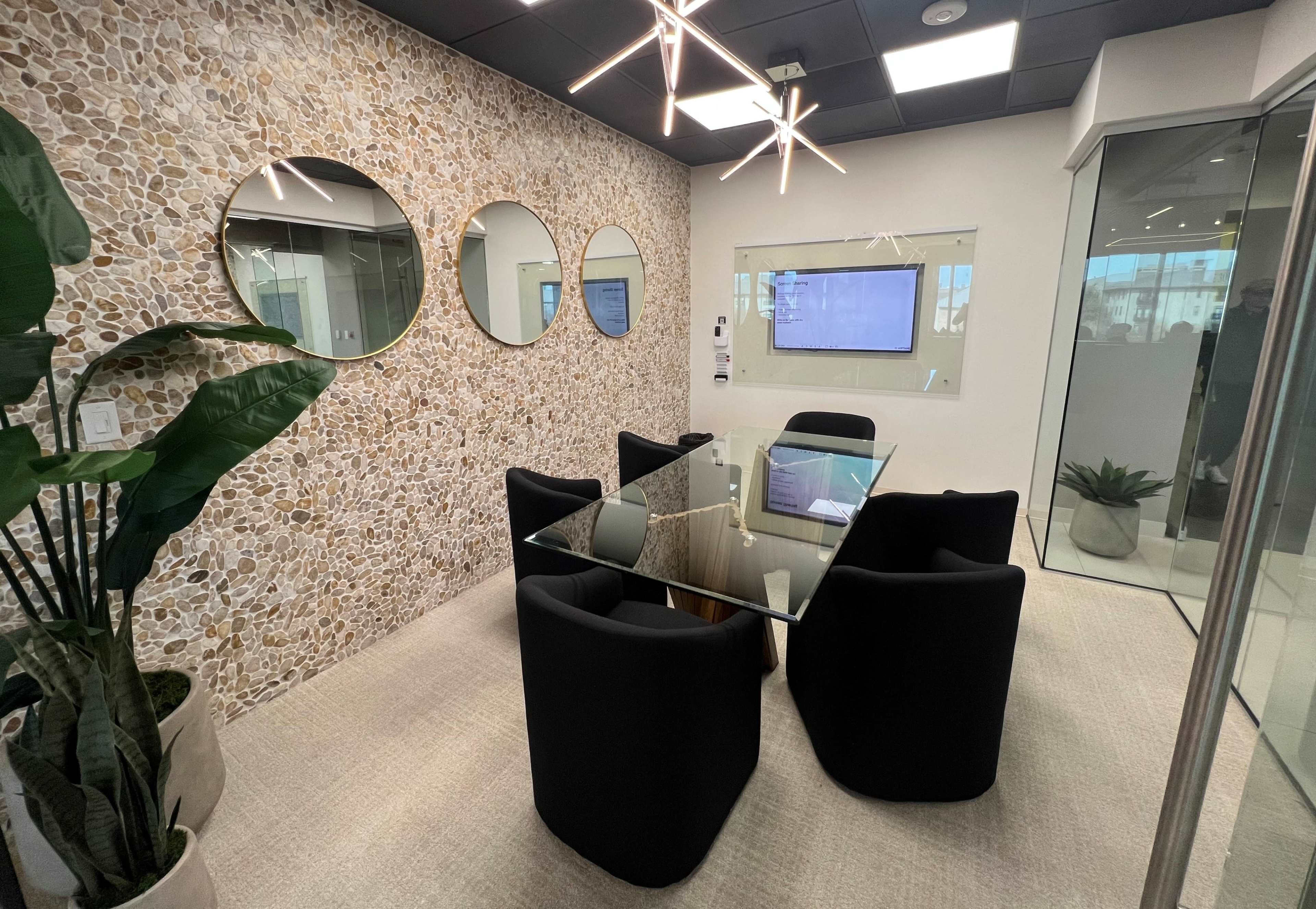 A modern conference room features a glass table surrounded by black chairs, with a stone wall and three round mirrors, along with a television and indoor plants.
