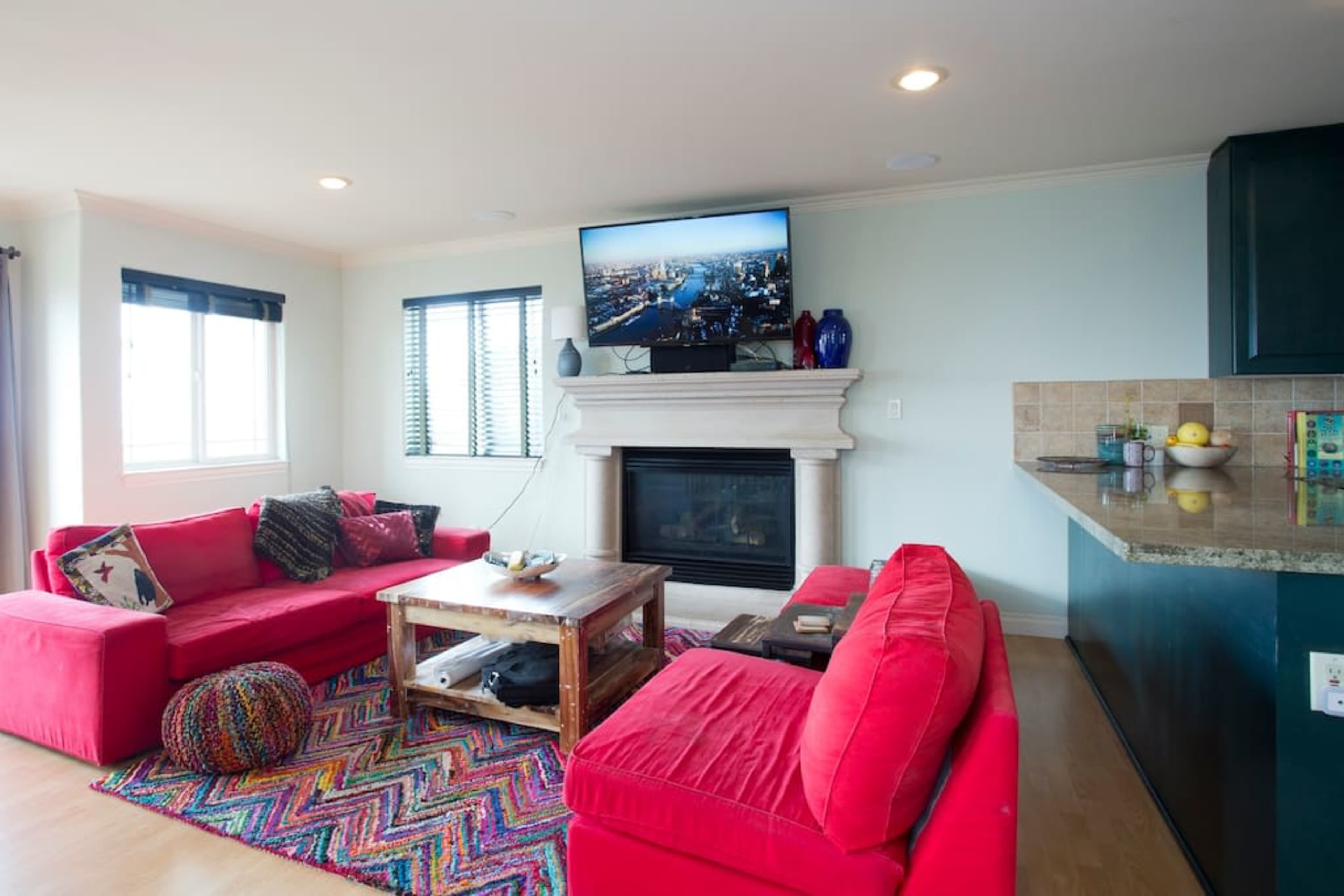 The living room features a red sofa set, a wooden coffee table, a fireplace with a television above it, and a vibrant area rug on the floor.