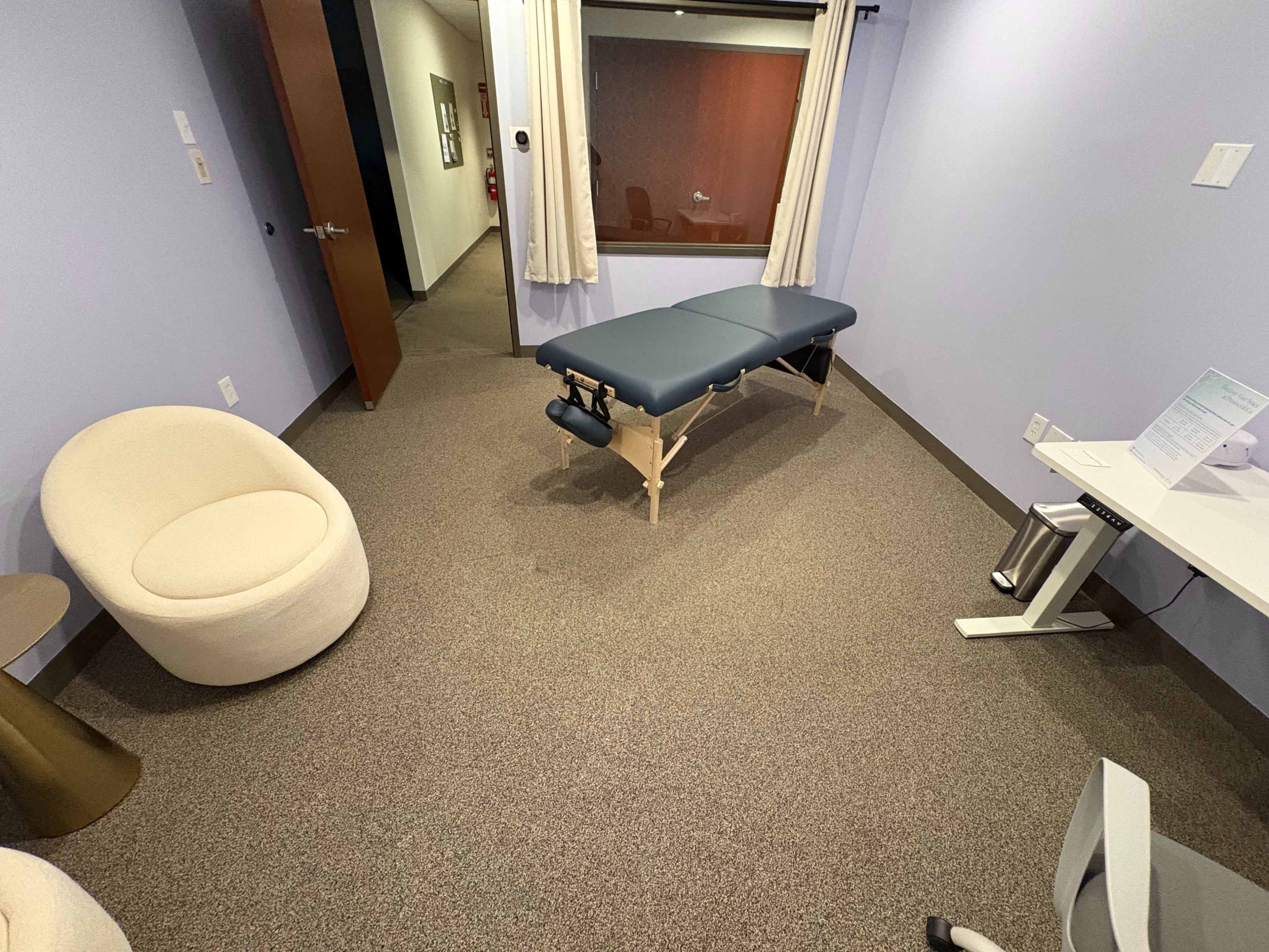 The image depicts a clinical room featuring a massage table, a round chair, a desk with a chair, and a large window.