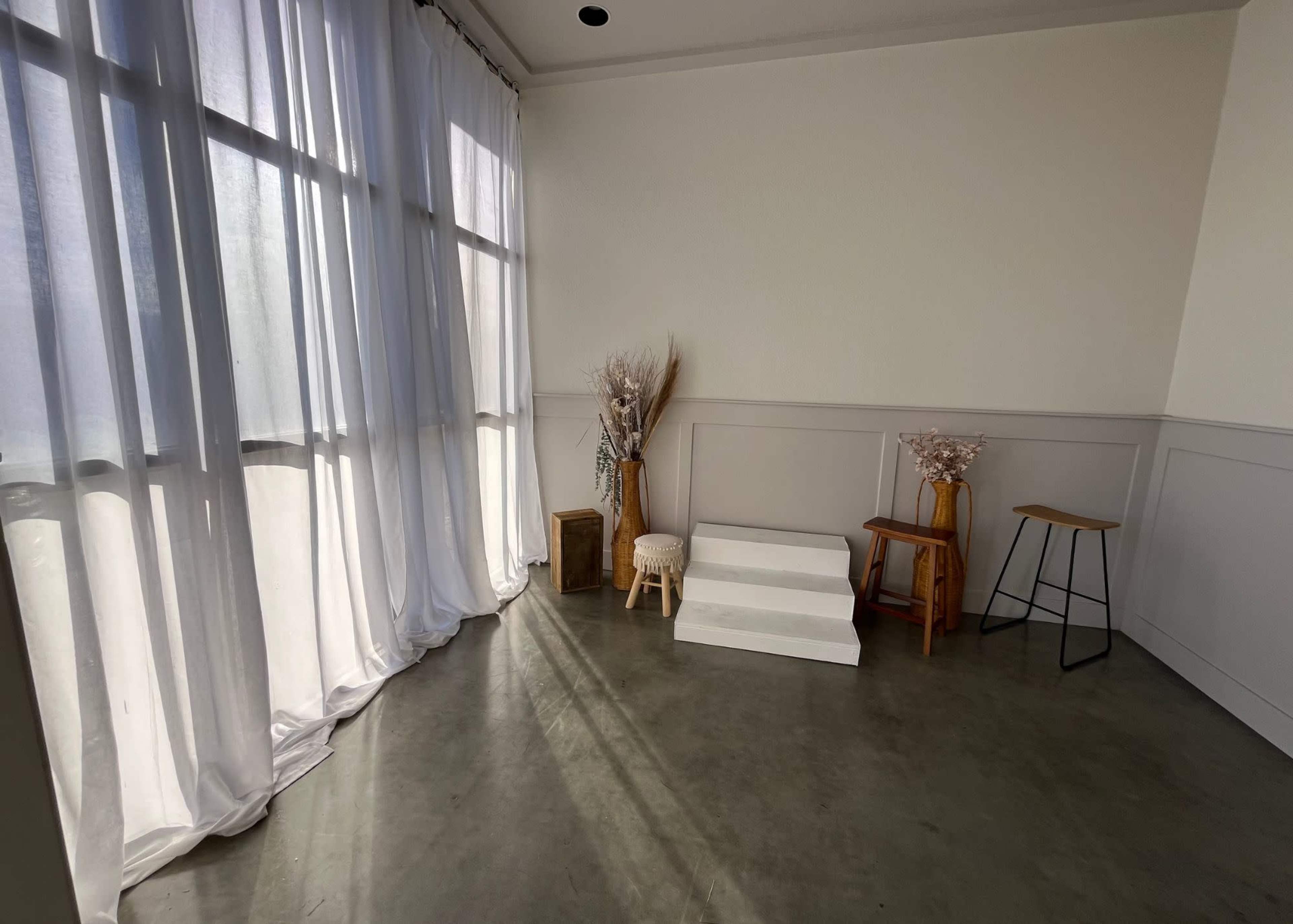 The image shows a bright, minimalistic interior space with large windows, sheer white curtains, and a few wooden furniture pieces arranged around a small set of white stairs.