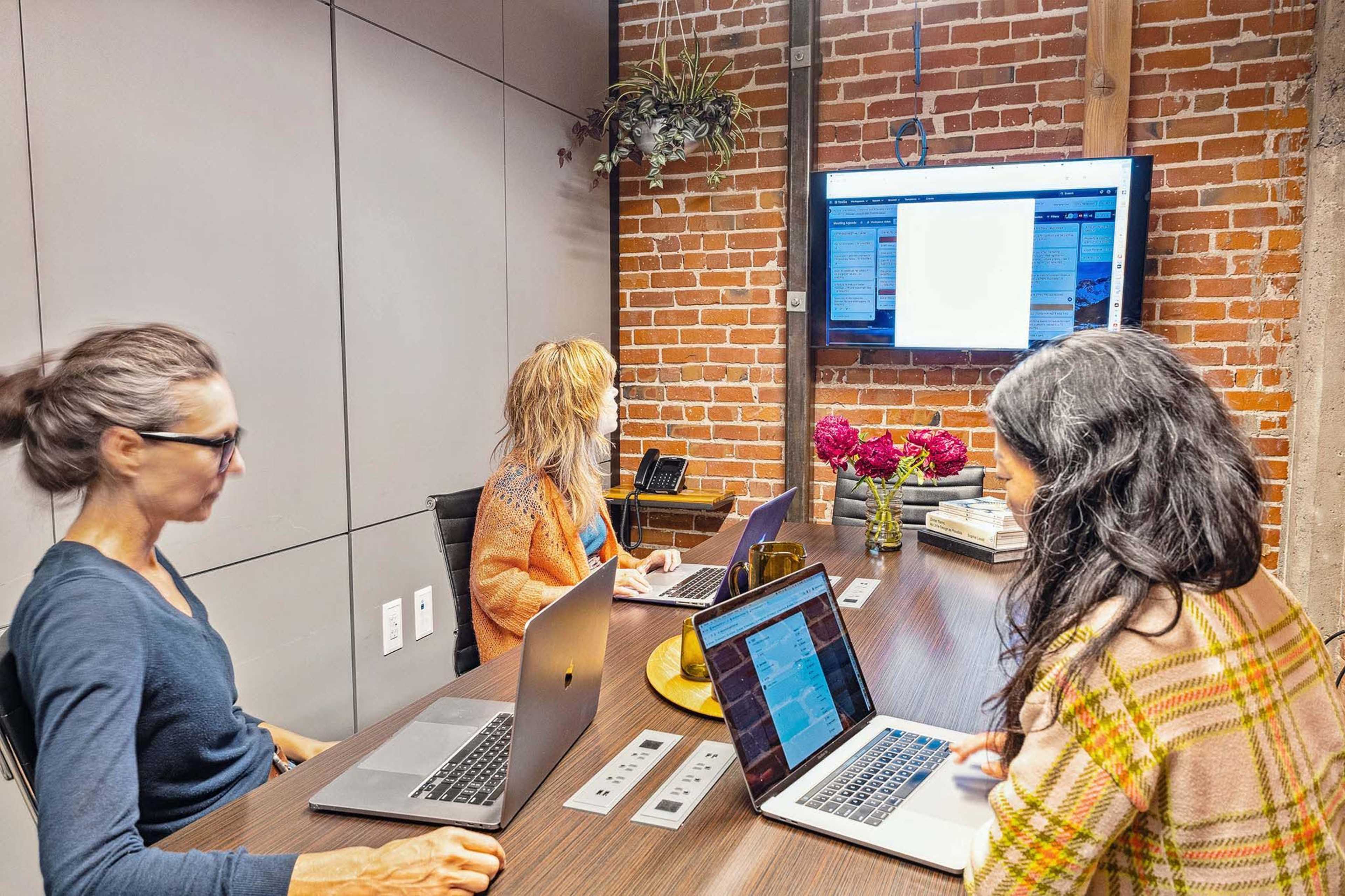 Three women work together in a conference room with a brick wall, laptops open in front of them, and a screen displaying documents or data.