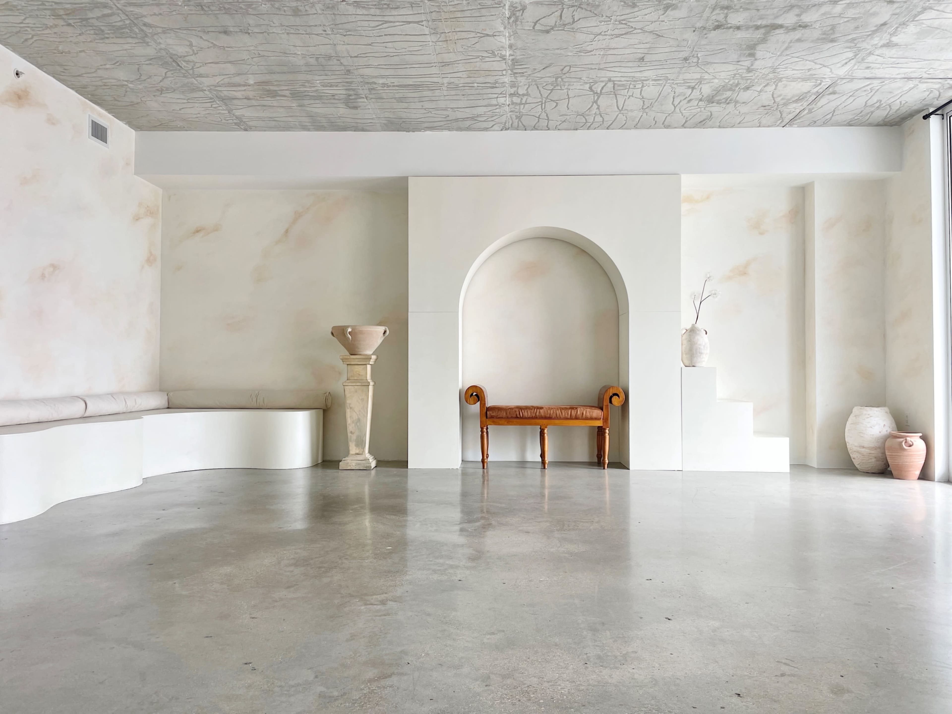 The image shows a minimalist interior space featuring a white wall nook with a sculptural vase, a wooden bench, a pedestal with a decorative urn, and a staircase leading to a raised area.