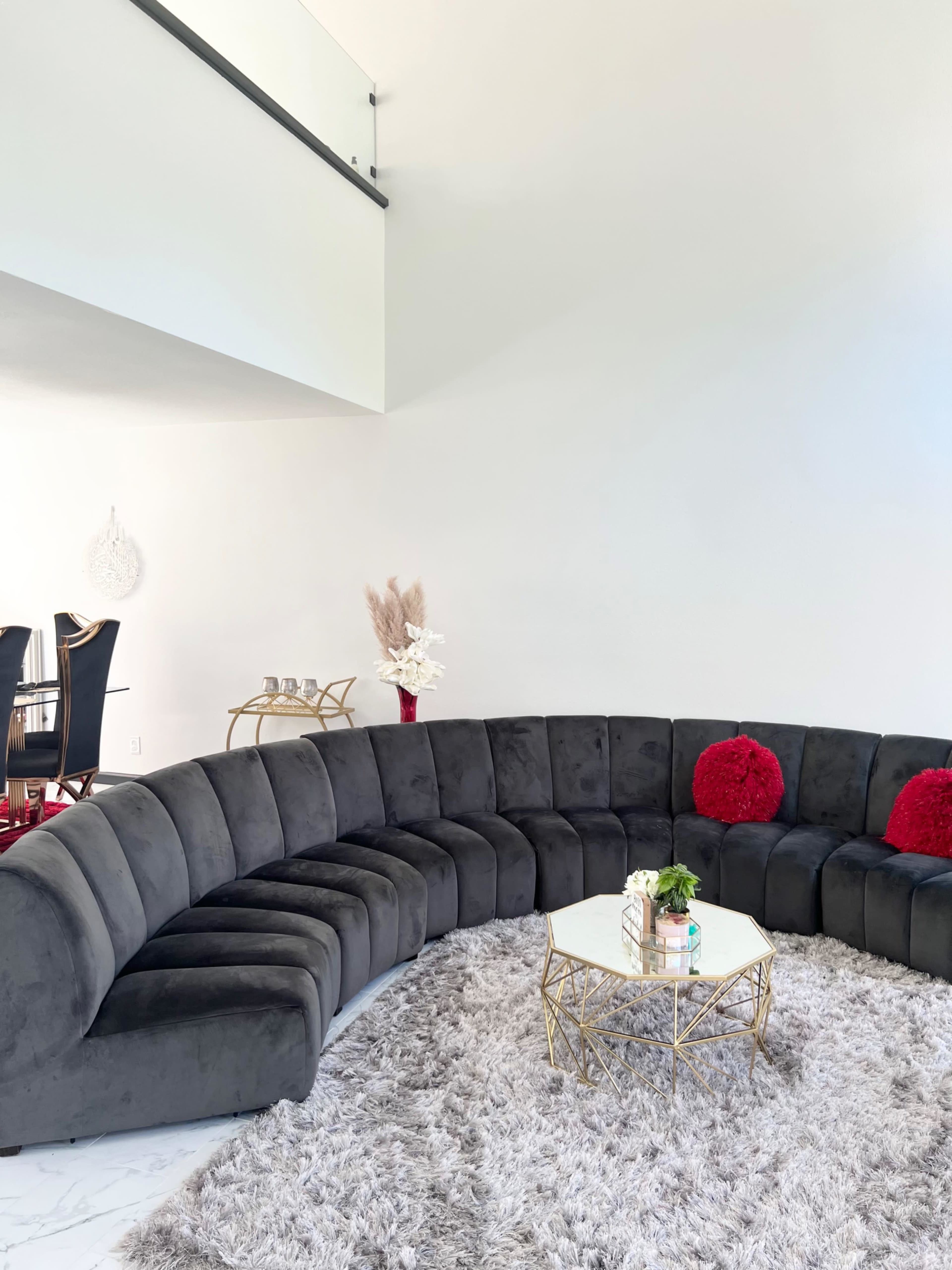 The image shows a modern, curved gray sectional sofa arranged around a glass coffee table, set on a fluffy rug in a bright room.