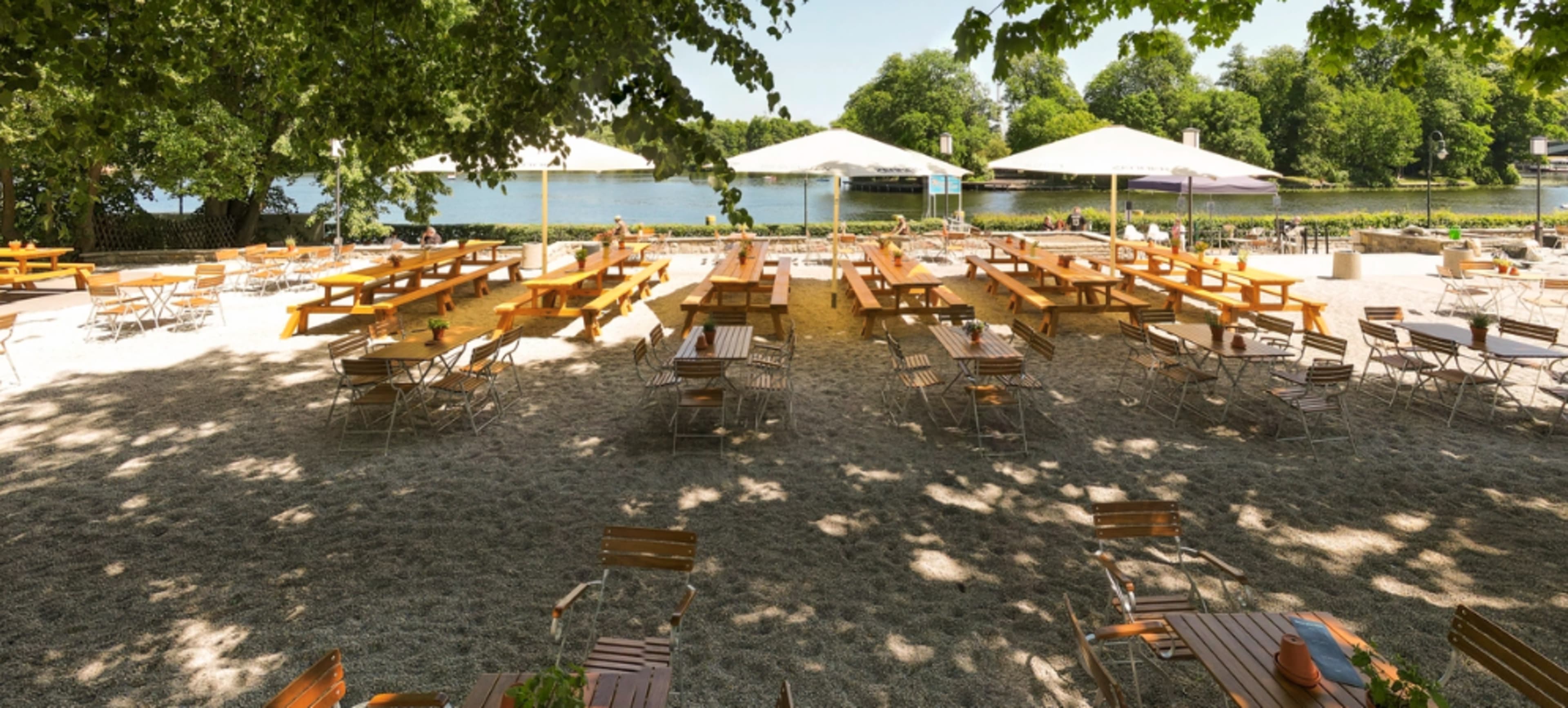 A sandy outdoor seating area with wooden tables and chairs under umbrellas, adjacent to a calm body of water and surrounded by trees.