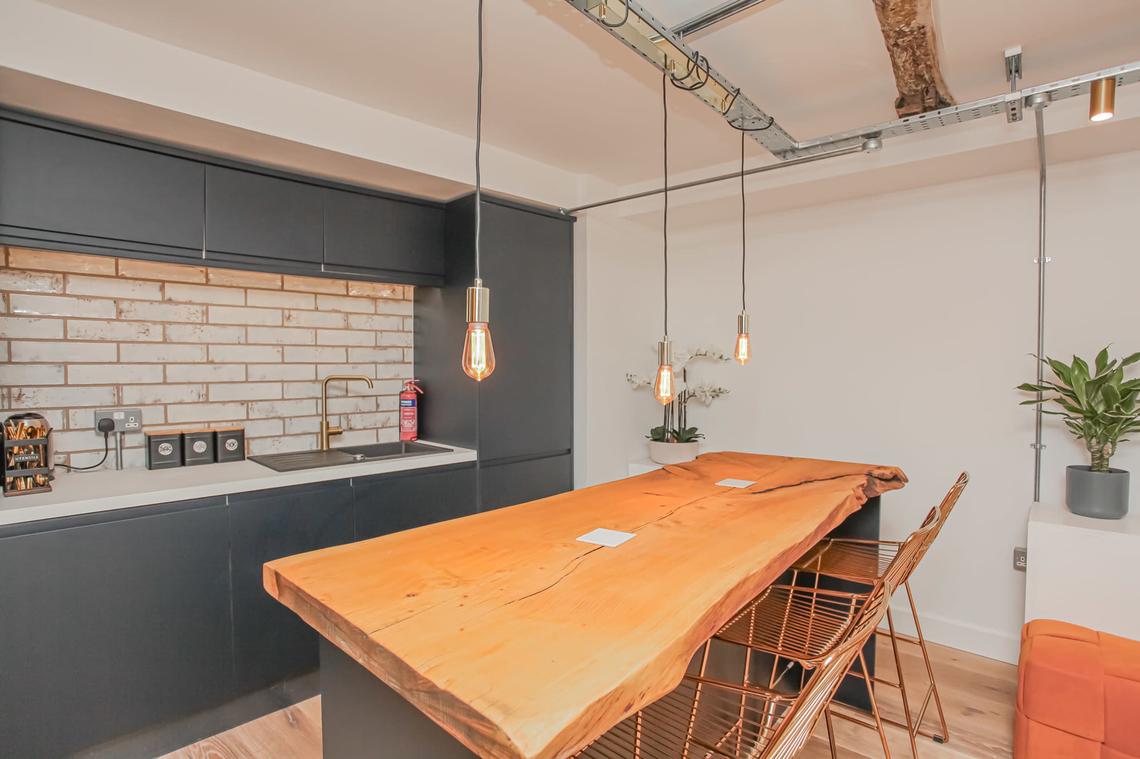 The image shows a modern kitchen featuring a wooden dining table with metal stools, a brick backsplash, and overhead pendant lights.