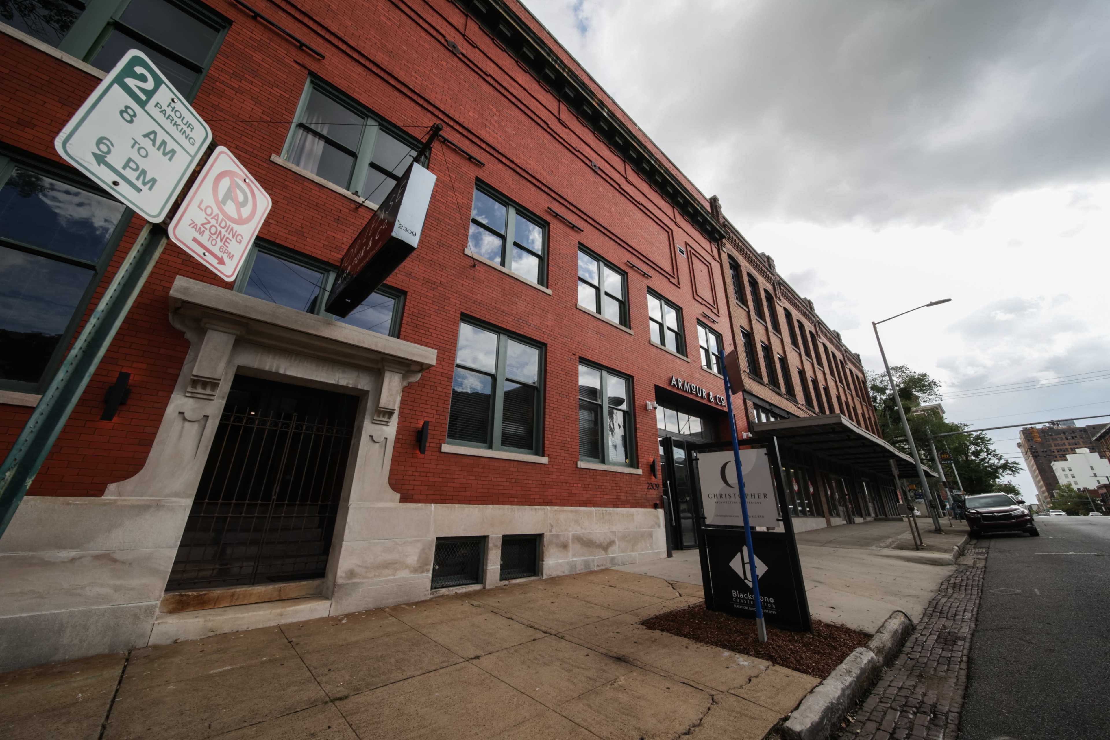 The image shows a red brick building with large windows and a stone entrance, situated on a street with a parking sign indicating restrictions.
