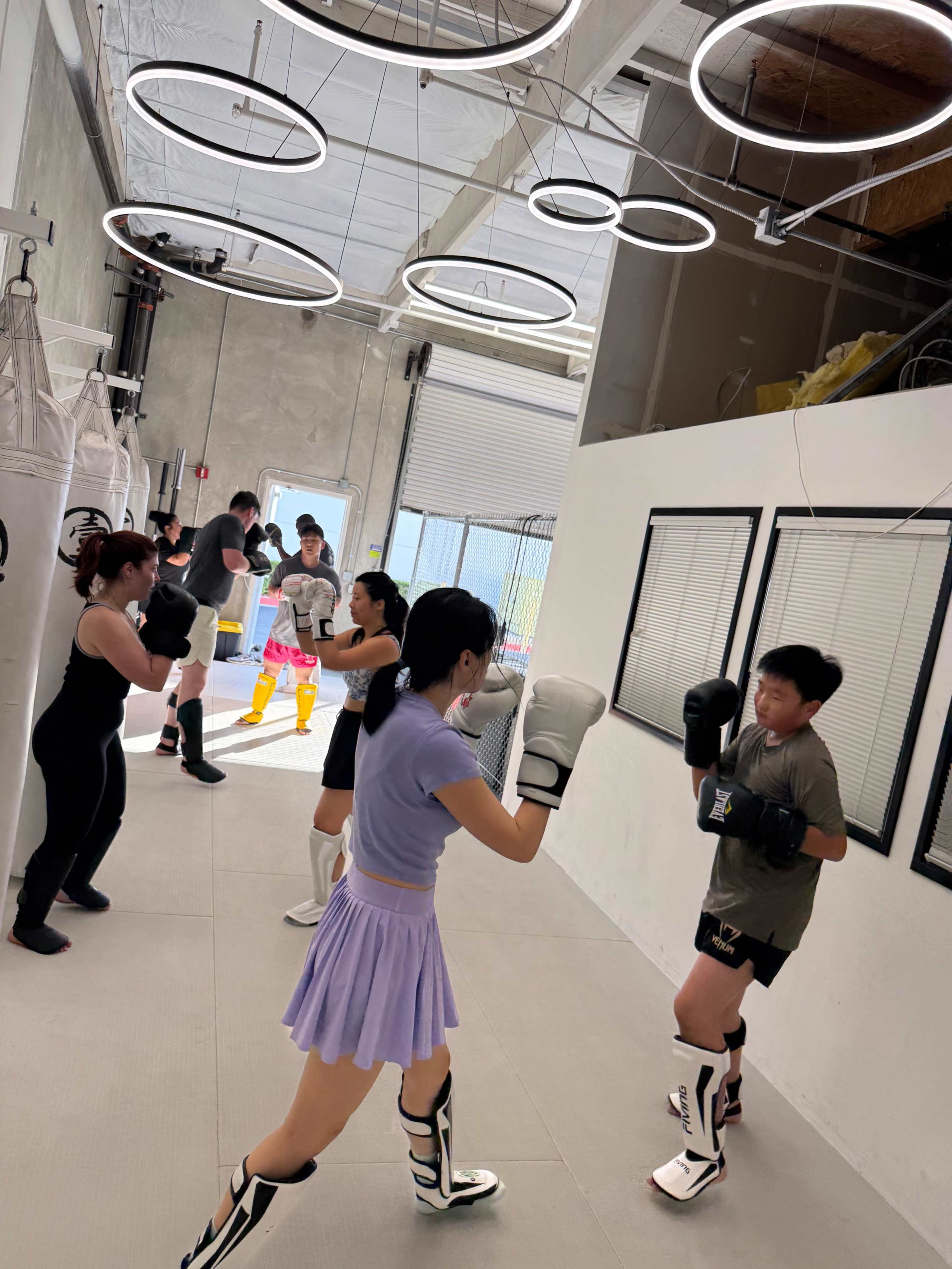A group of individuals is engaged in various boxing exercises inside a modern gym with punching bags and decorative lighting.