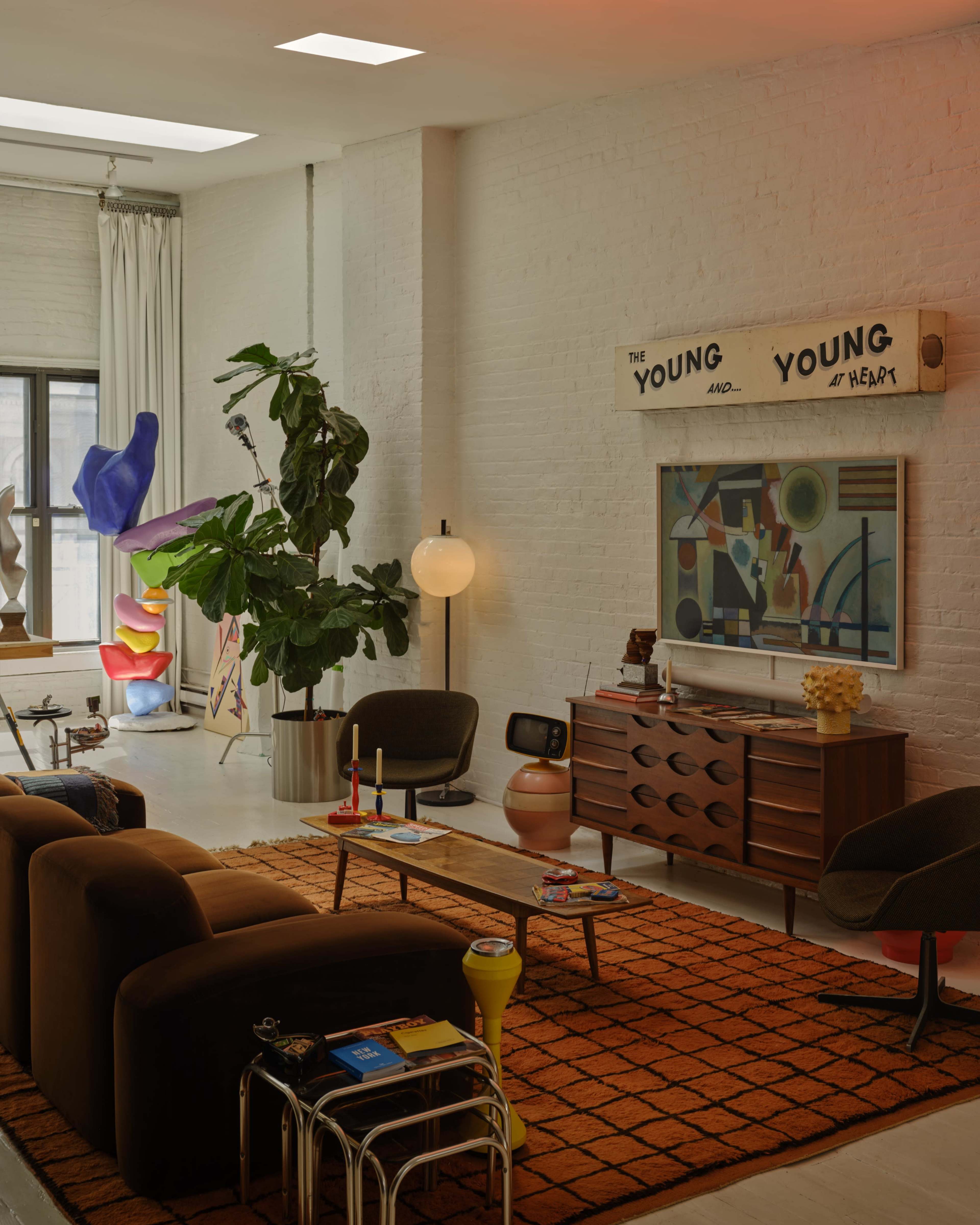 The image shows a modern living room featuring two brown chairs, a wooden coffee table, a colorful rug, a large plant, and various art pieces on the walls.