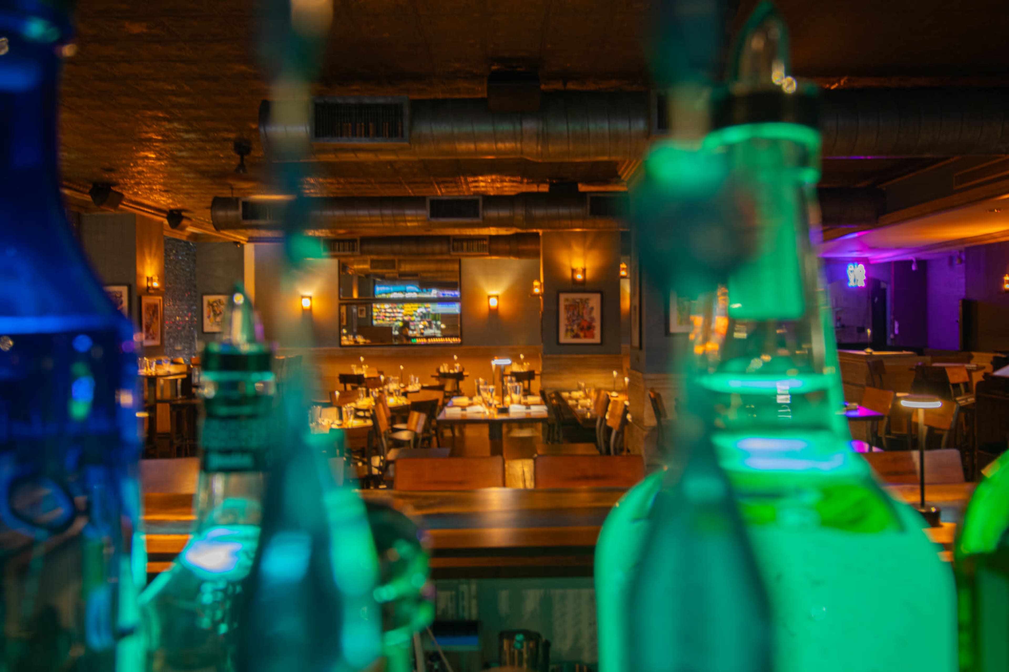 The image shows a restaurant interior with tables set for dining, viewed through colorful glass bottles in the foreground.