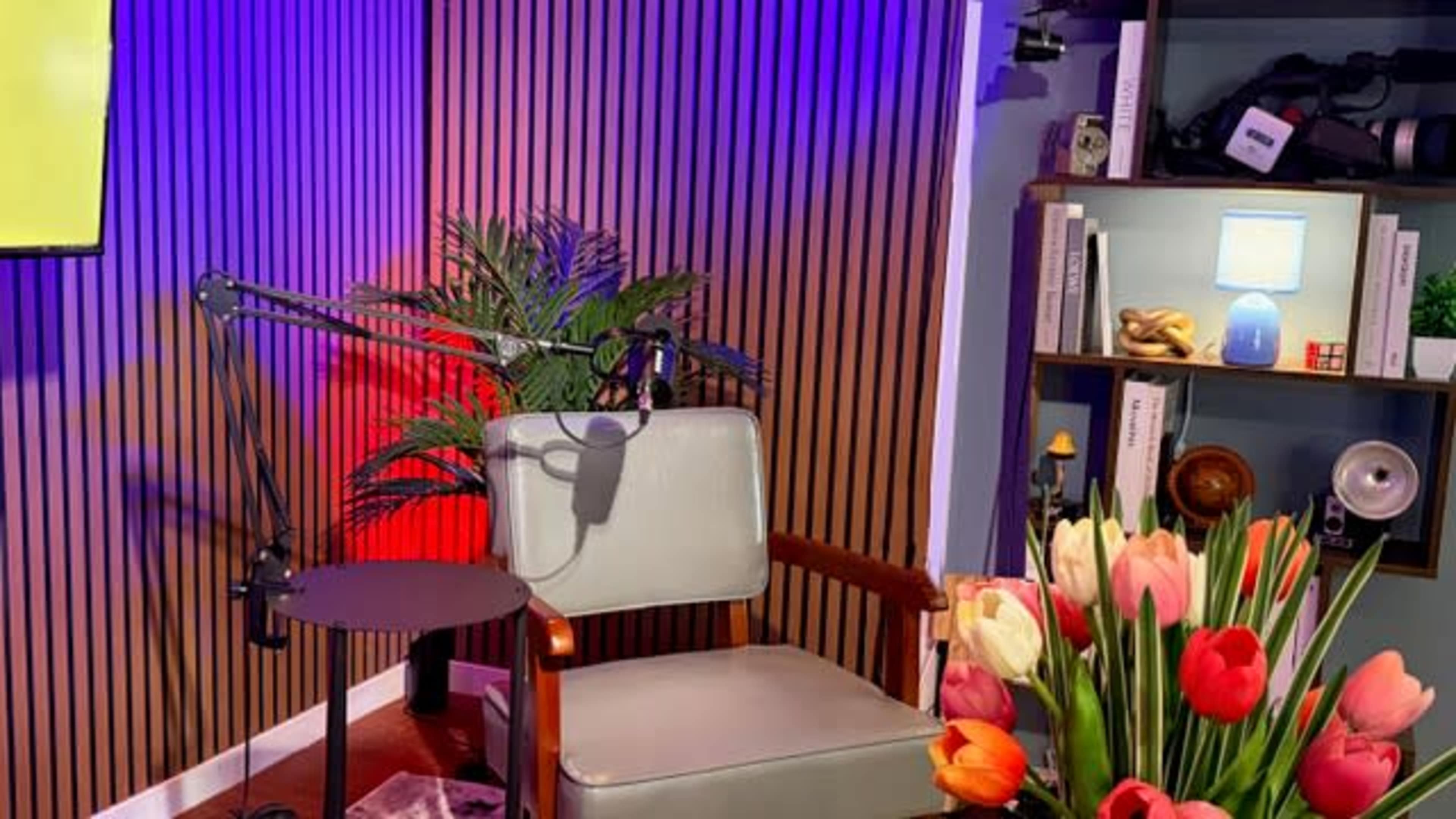 The image shows a modern interview setup with a single chair, a small round table, a microphone on an arm, vibrant flowers, and decorative shelves in the background.
