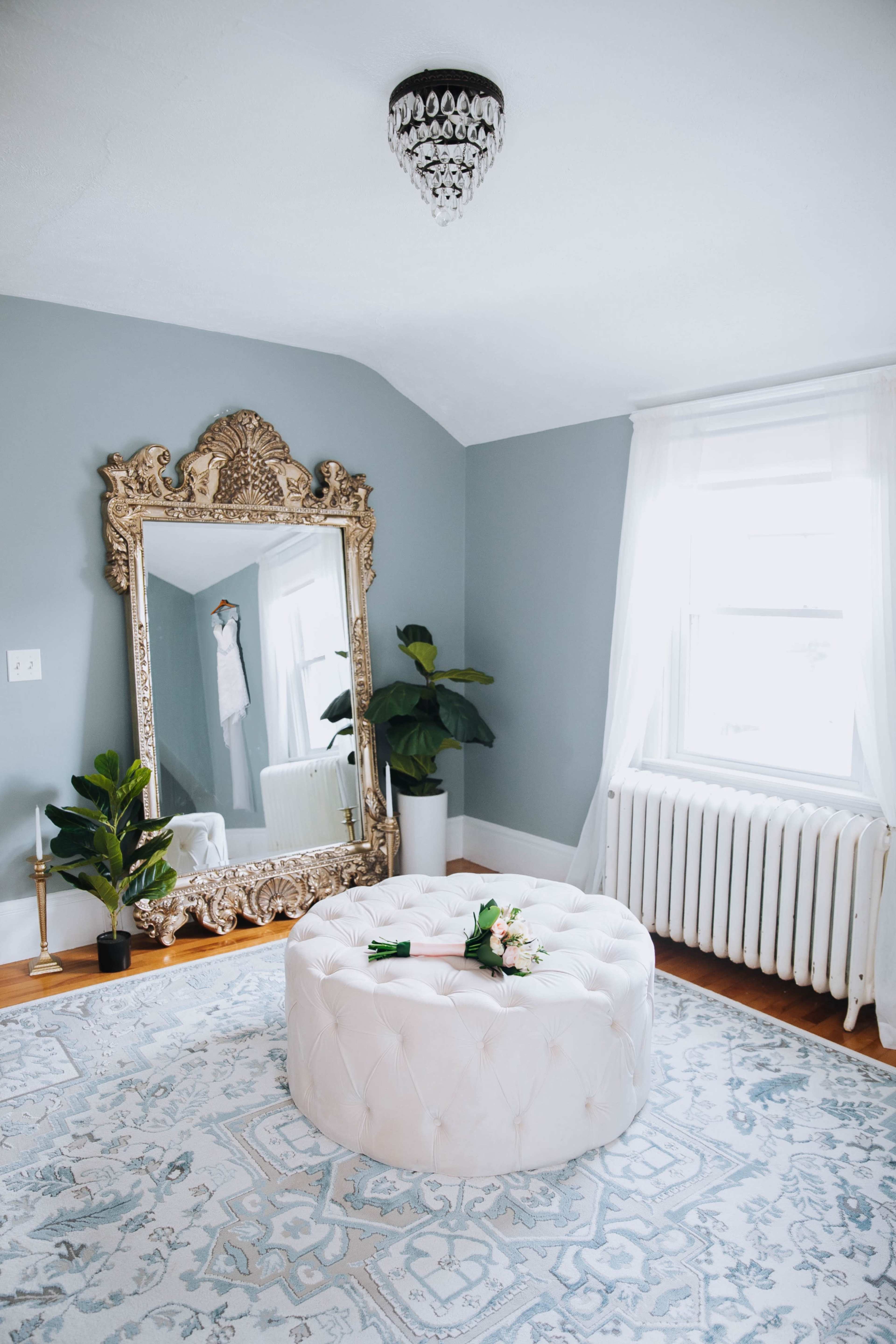 The room features a large ornate mirror, a tufted white ottoman, and a potted plant, all set against a light blue wall with a window letting in natural light.
