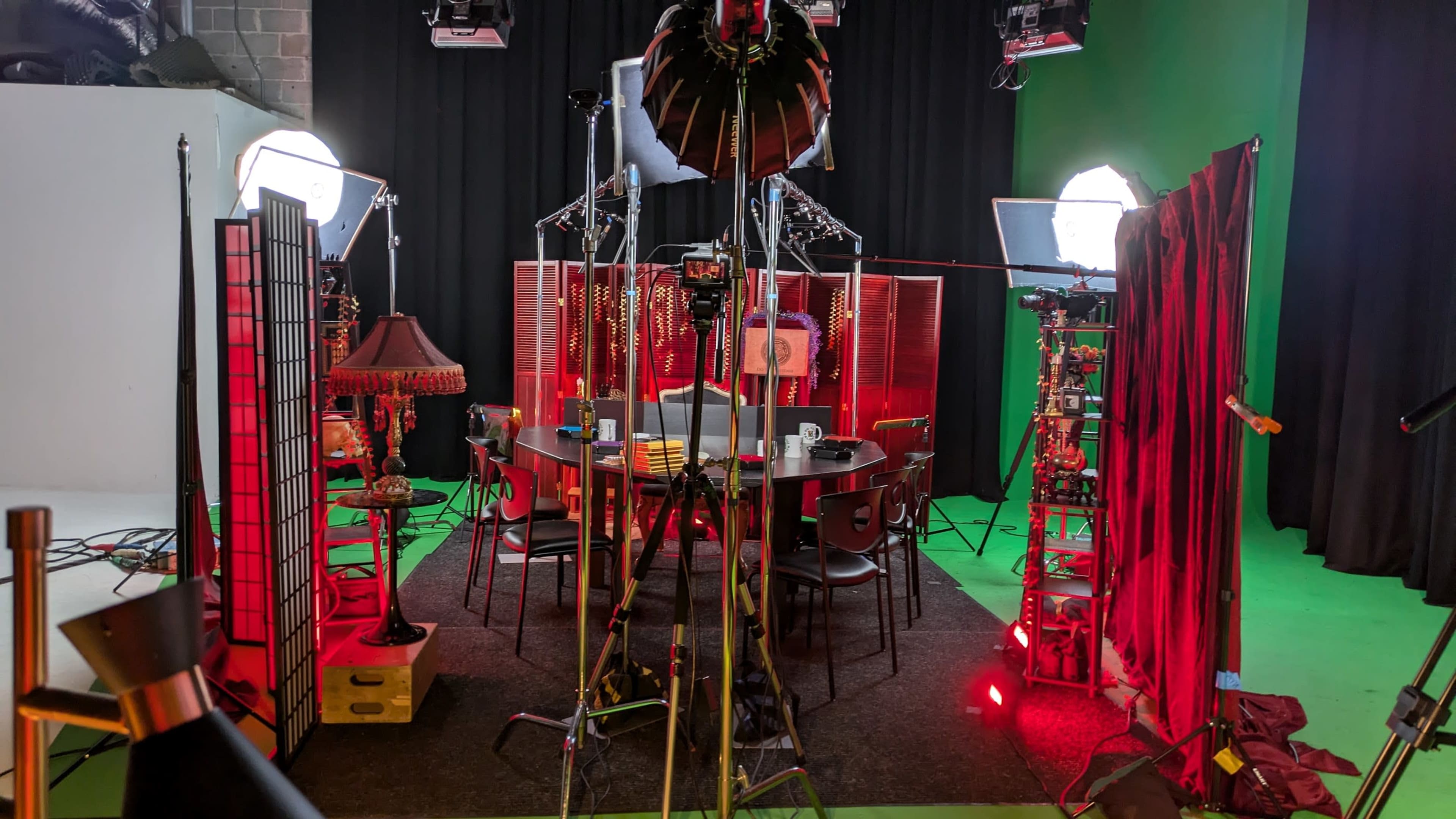 The image shows a studio set with a round table surrounded by chairs, illuminated by various lights and flanked by decorative screens.