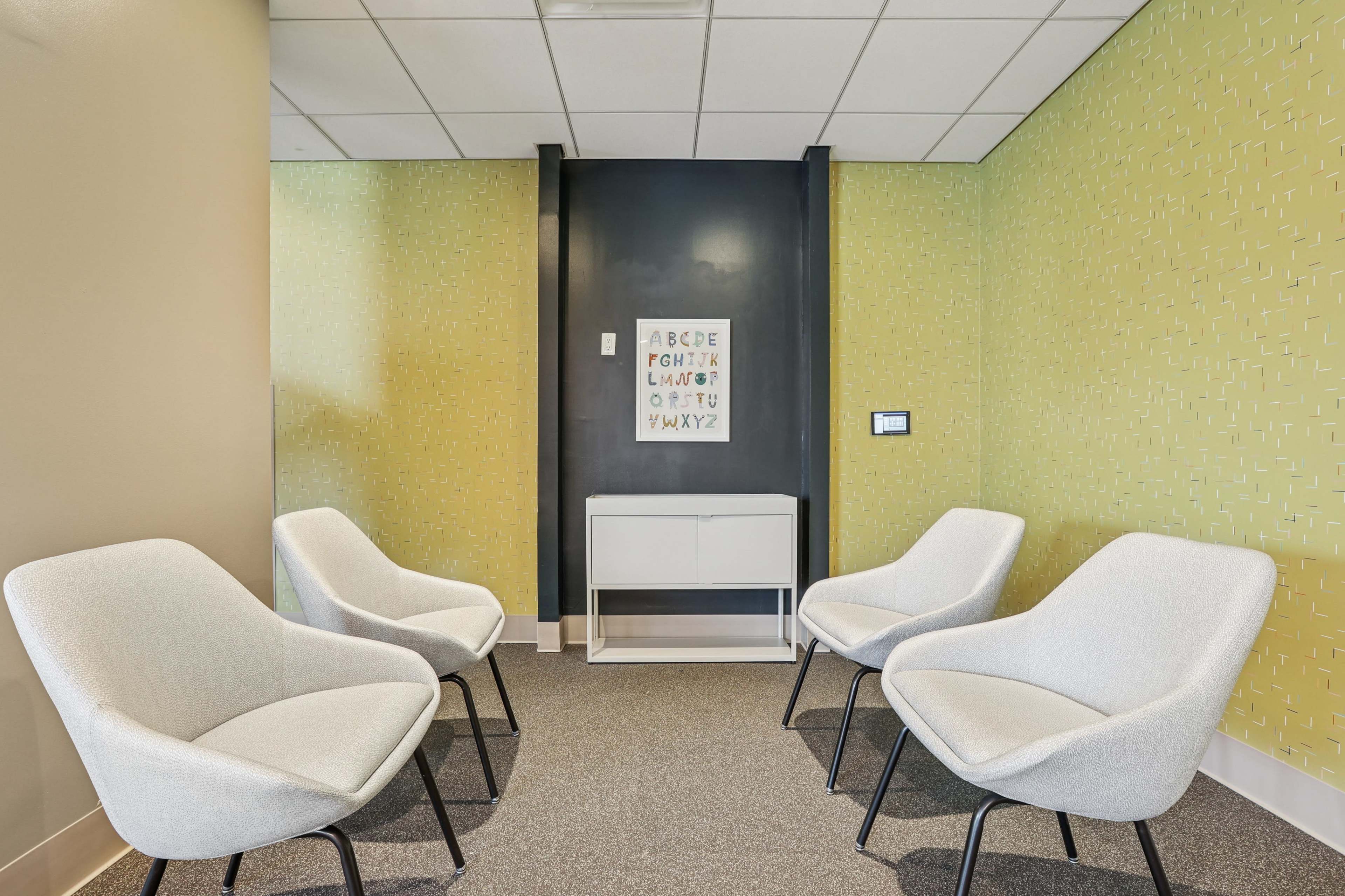 The image shows a small waiting area with four white chairs arranged around a white cabinet against a green patterned wall.