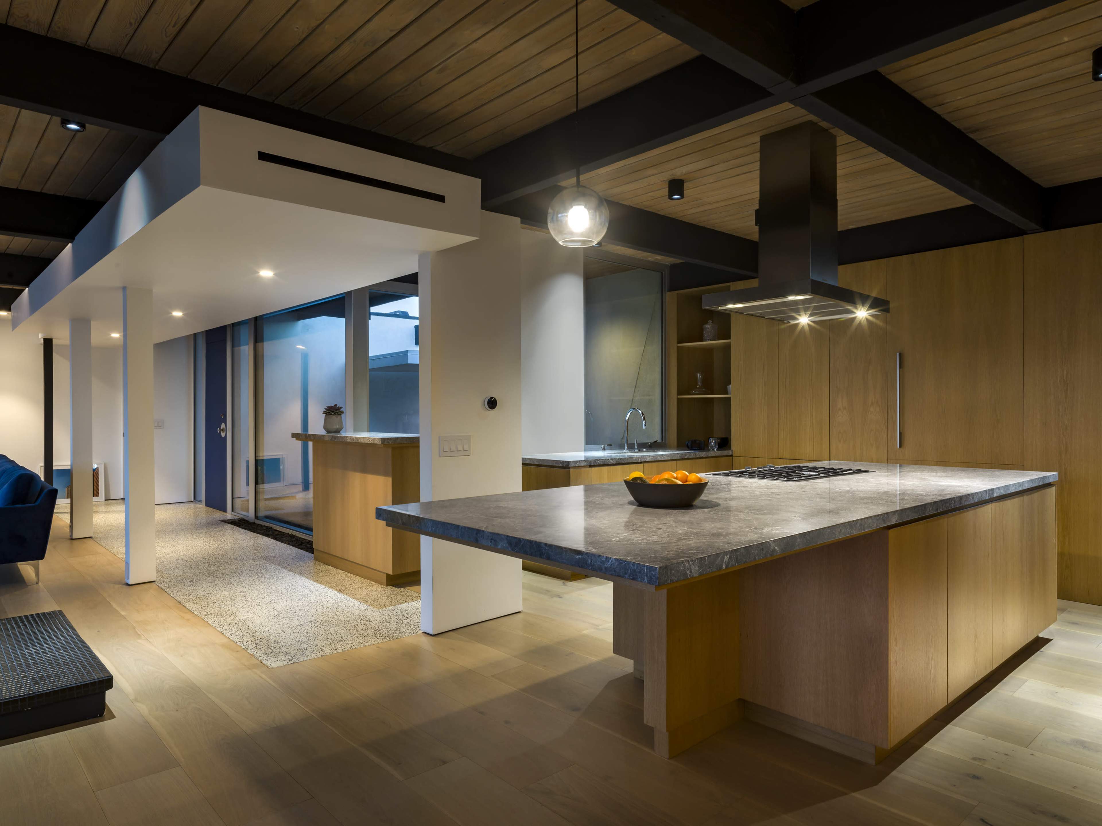 The image shows a modern kitchen with wooden cabinetry, a large island featuring a stove, and an open layout that connects to a well-lit living area.