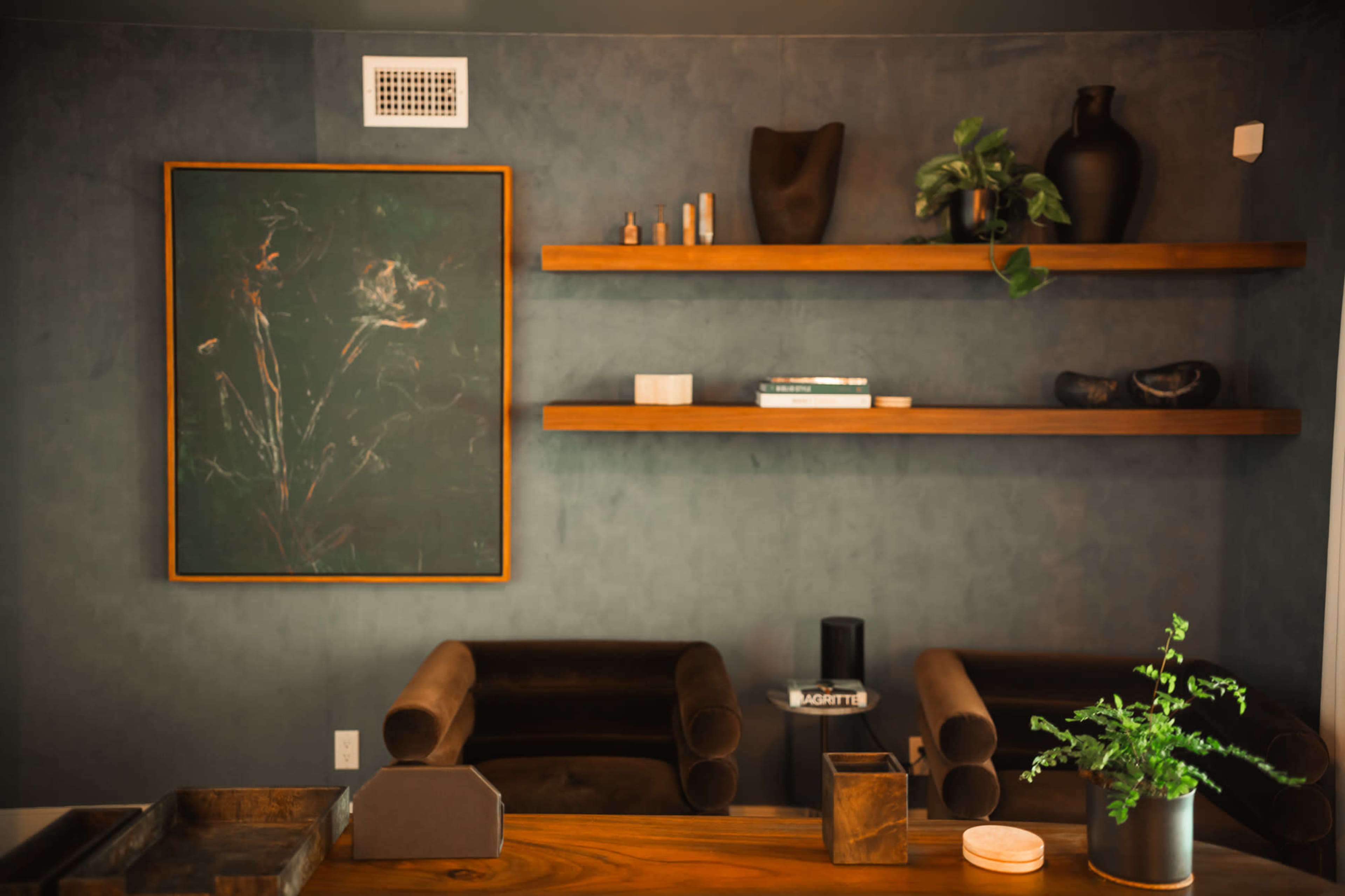 A styled interior features a dark wall with a large painting, wooden shelves displaying various objects, and two brown armchairs.