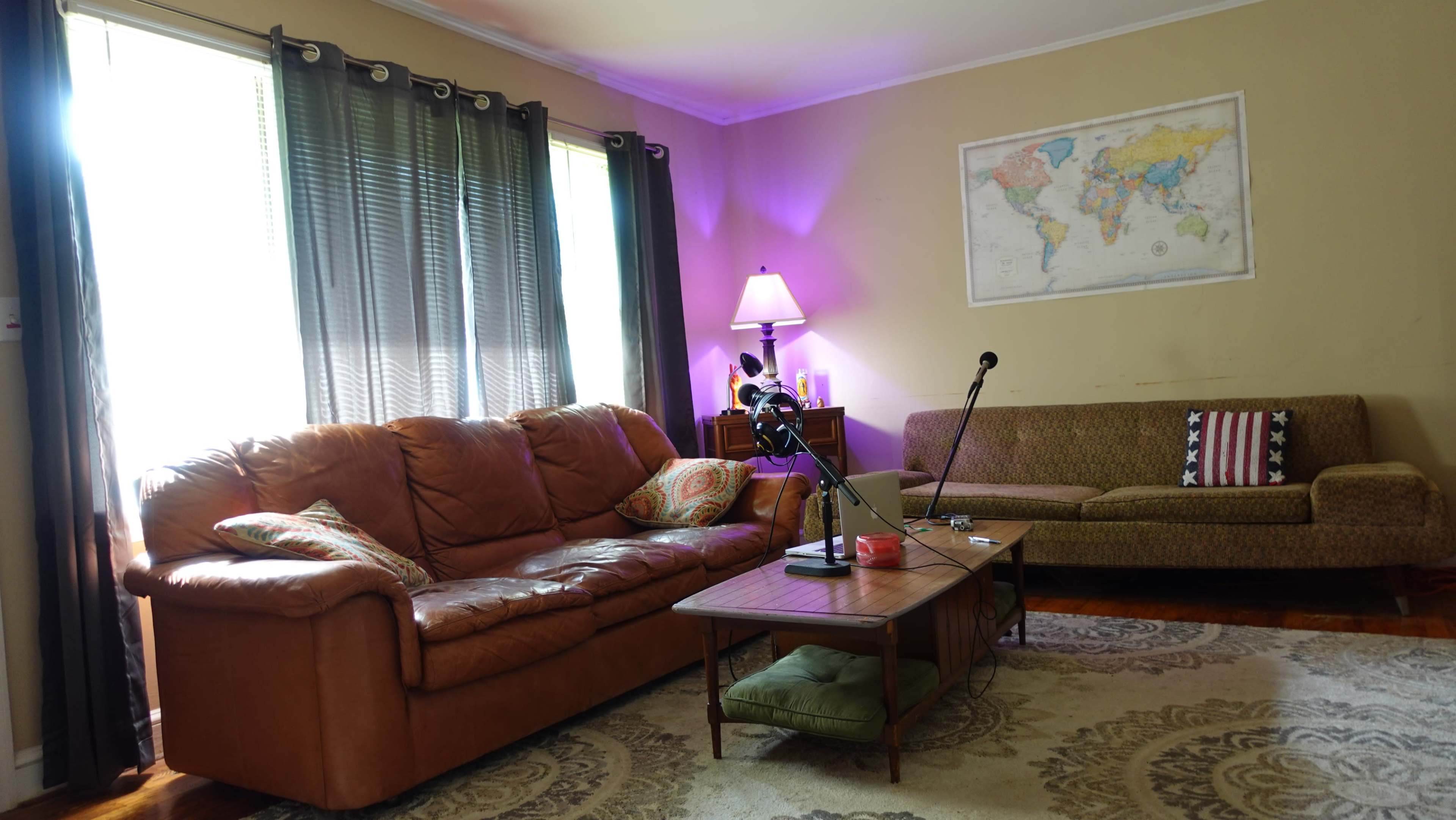 A living room features a brown leather sofa, a green couch, a wooden coffee table with a microphone setup, and a world map on the wall.