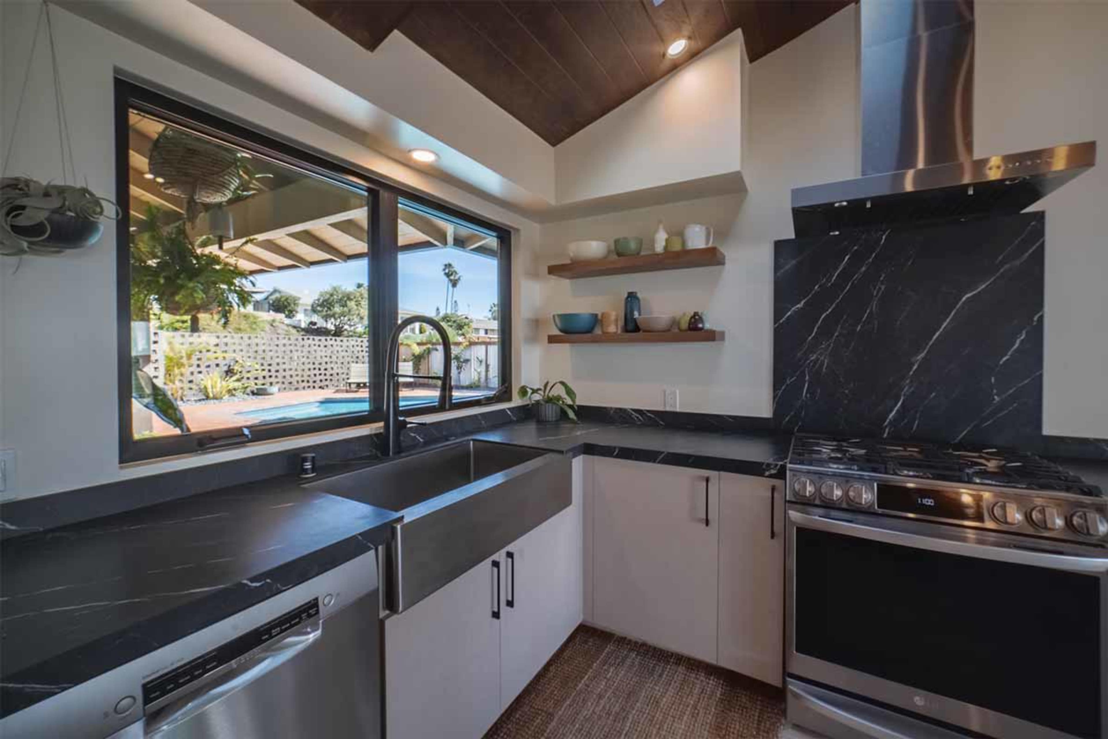 A modern kitchen features dark marble countertops, stainless steel appliances, and a large window overlooking a pool area.