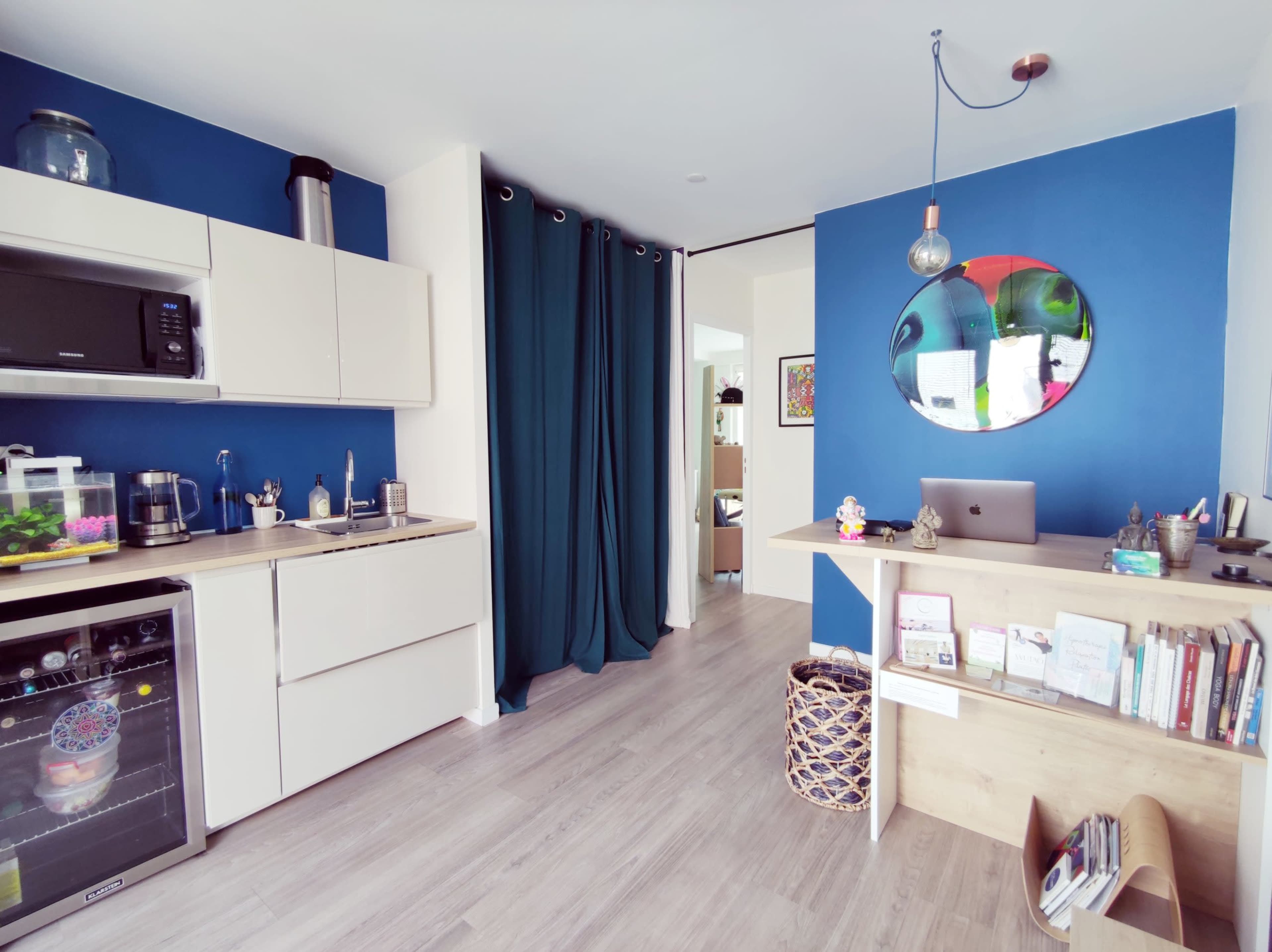 The image shows a modern kitchen with beige cabinetry and a blue accent wall, featuring a dining area with a small desk and shelves.