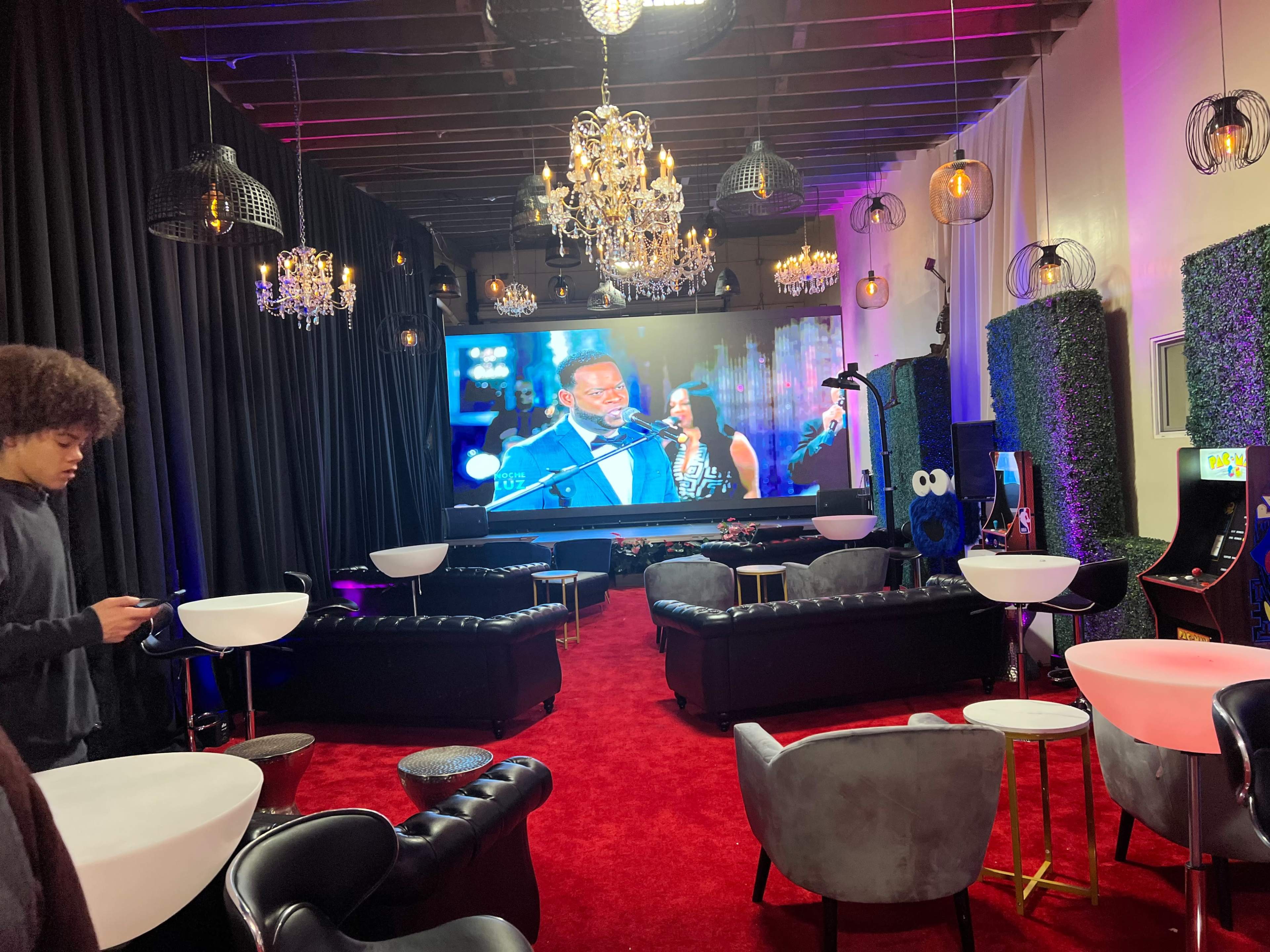 The image shows a stylish lounge area with a large screen displaying a performance, surrounded by plush seating, decorative lighting, and arcade machines.