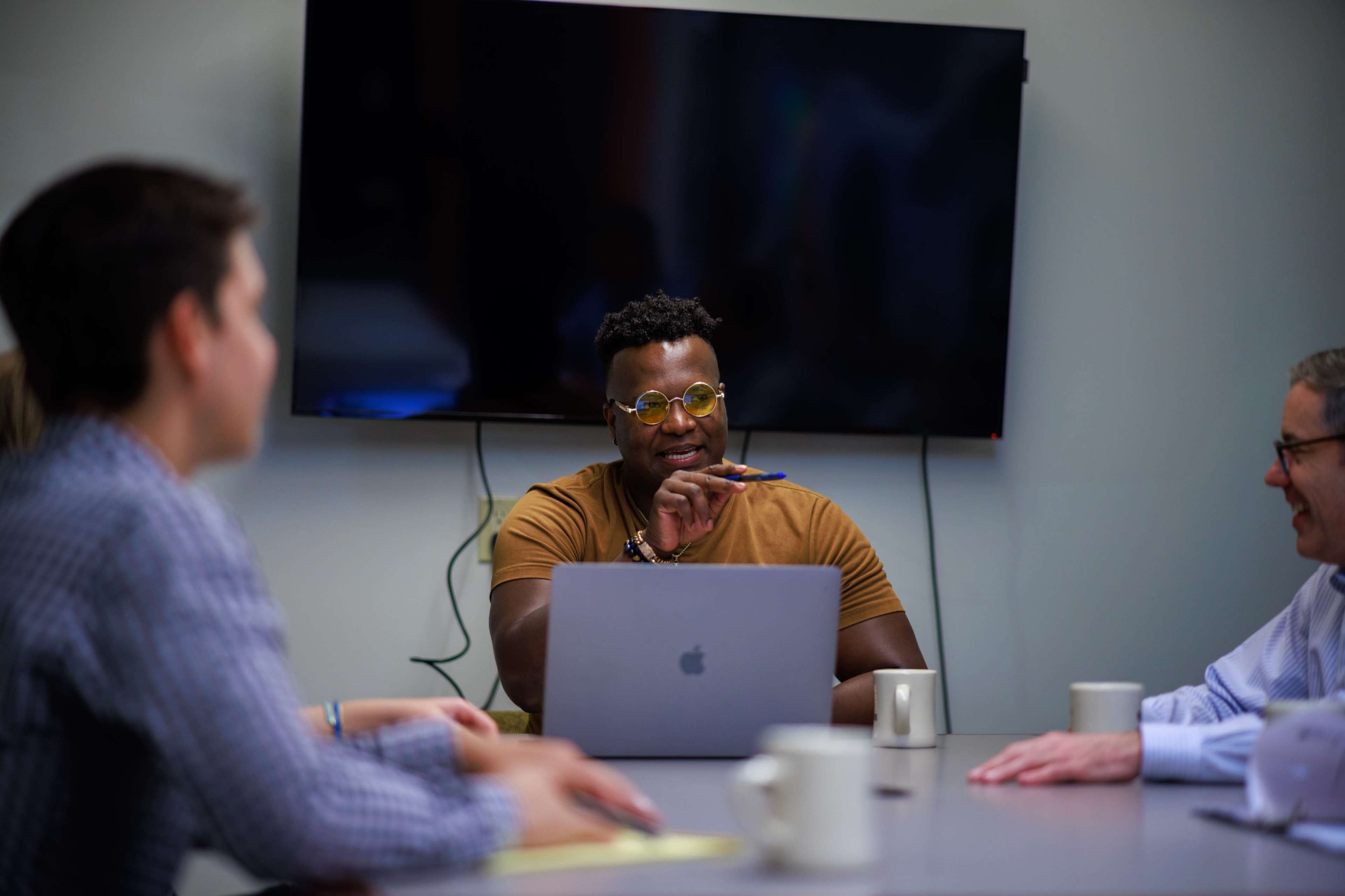 A person with glasses is sitting at a conference table, using a laptop while engaging in a discussion with three others.