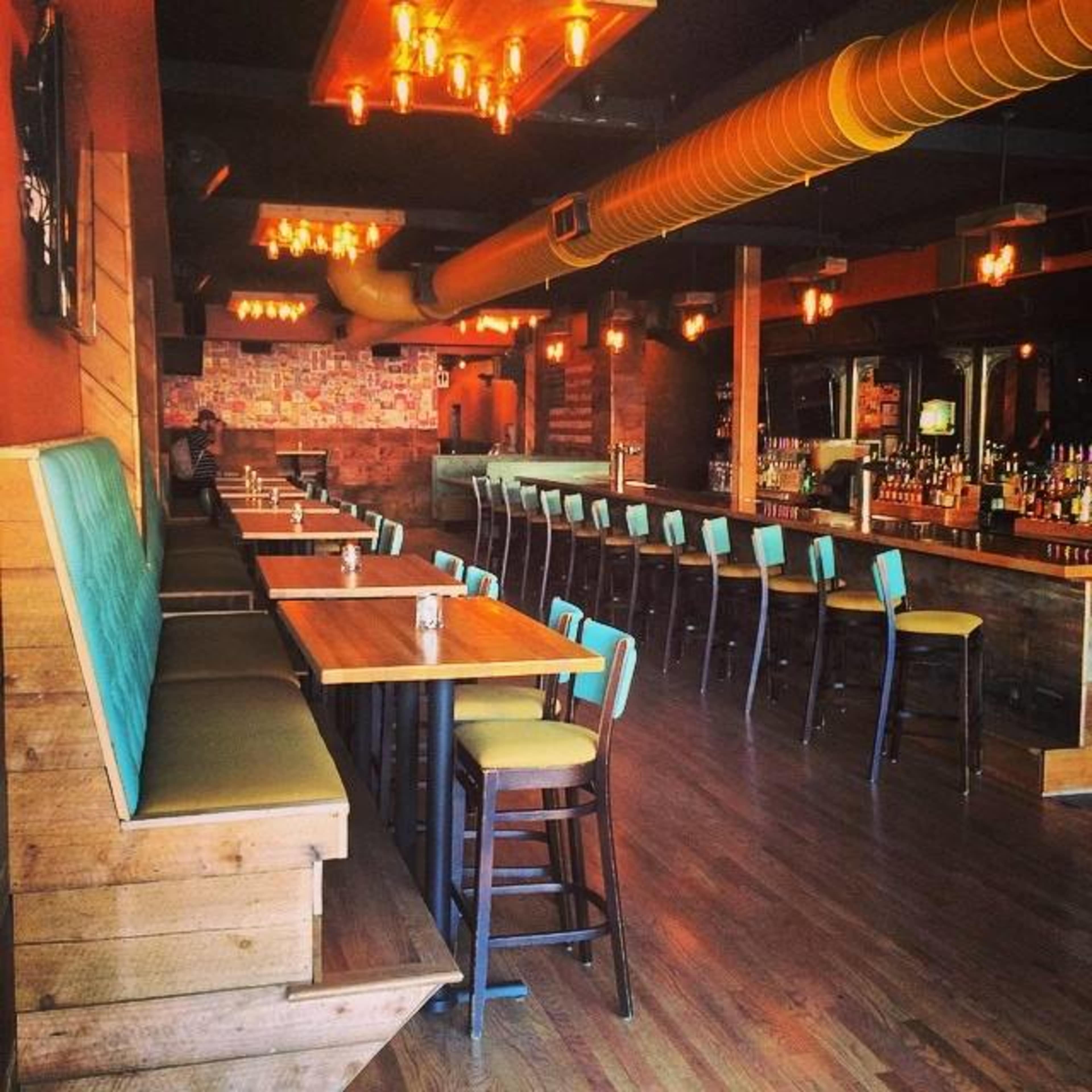 The image shows an empty restaurant interior with wooden tables, turquoise bar stools, and warm lighting fixtures.