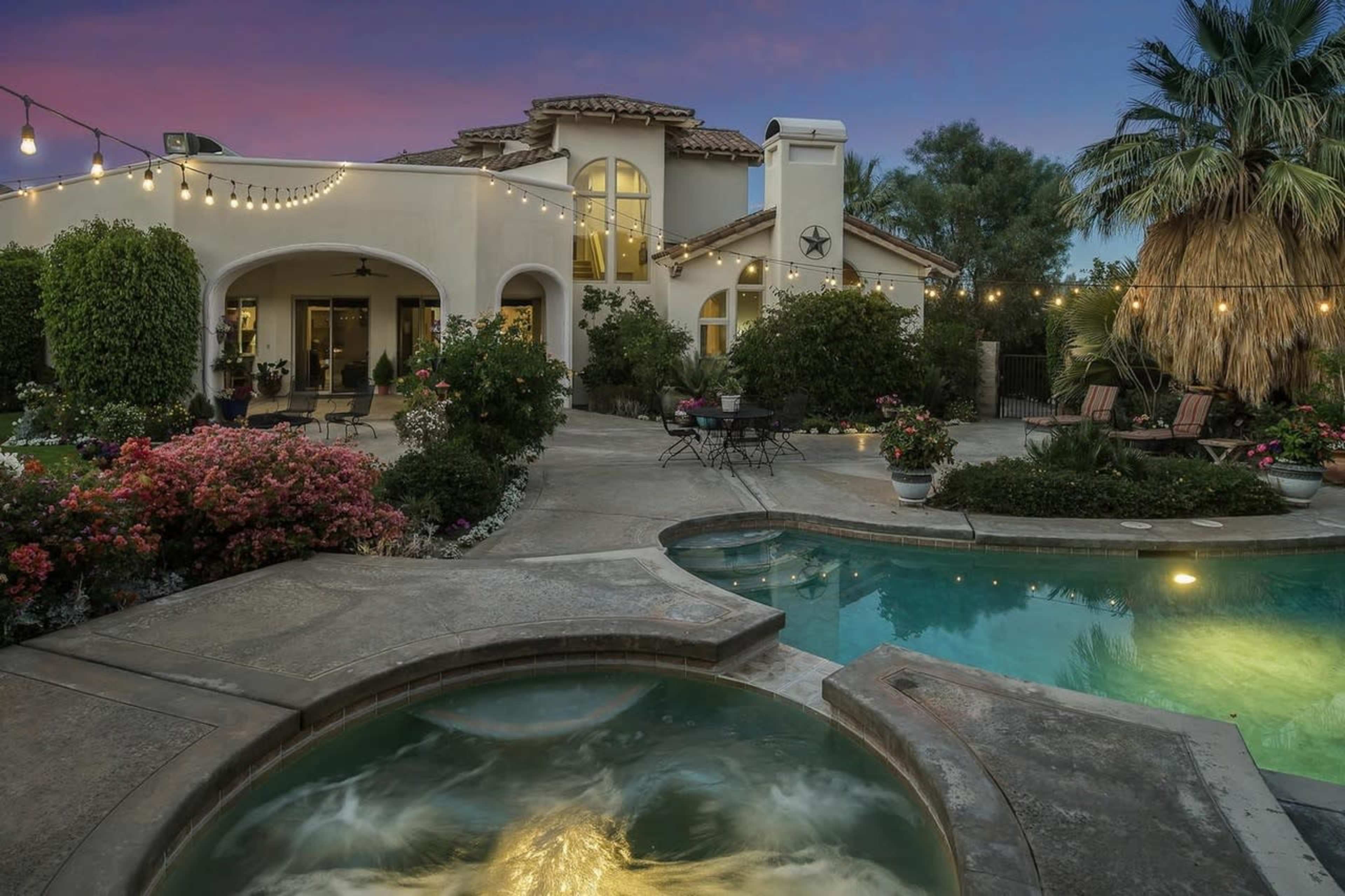 A well-lit backyard features a pool and spa surrounded by vibrant landscaping and a charming home in the background.