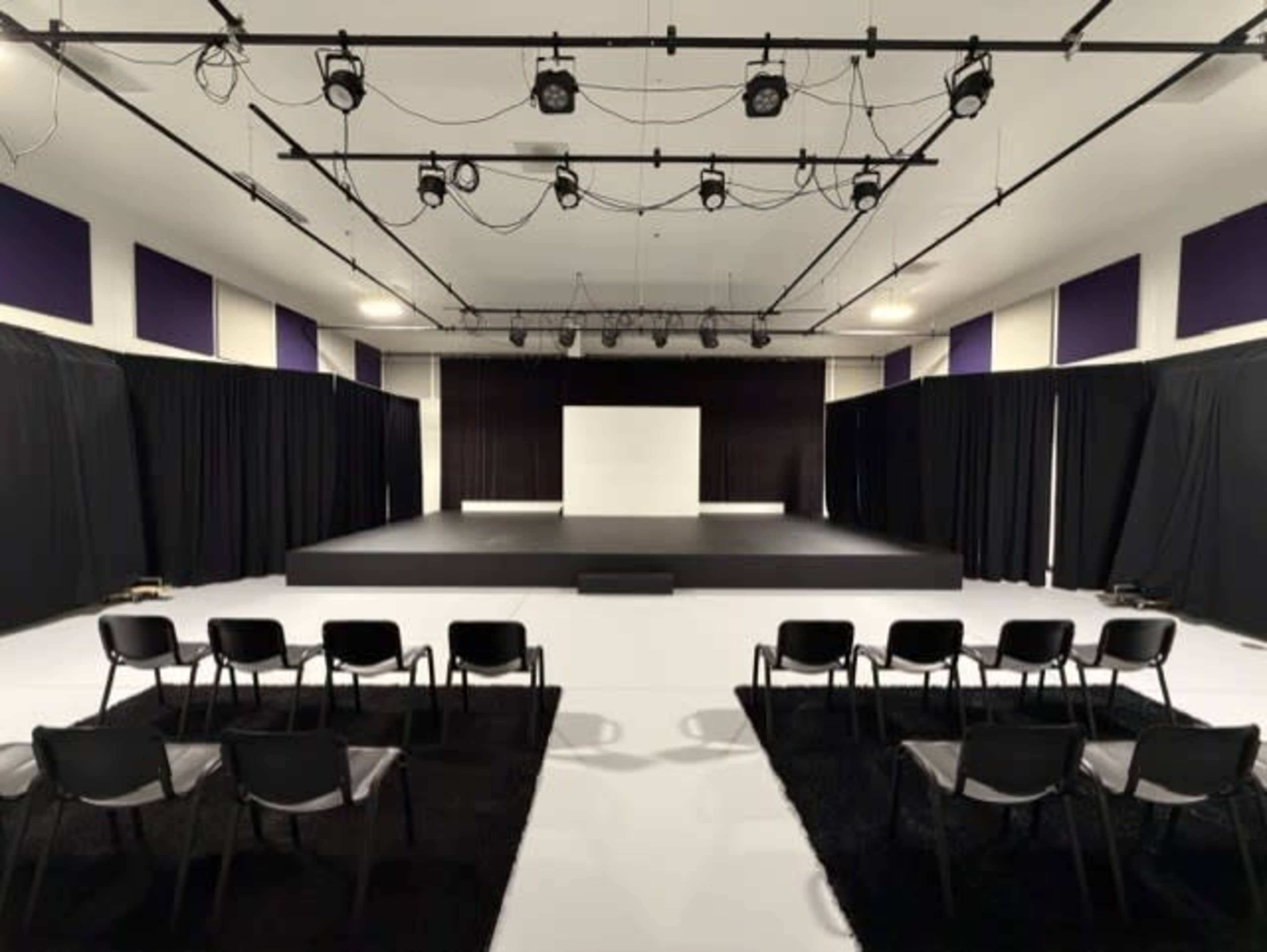 The image shows a theater stage setup with a large black platform, surrounded by black curtains and rows of chairs facing the stage.