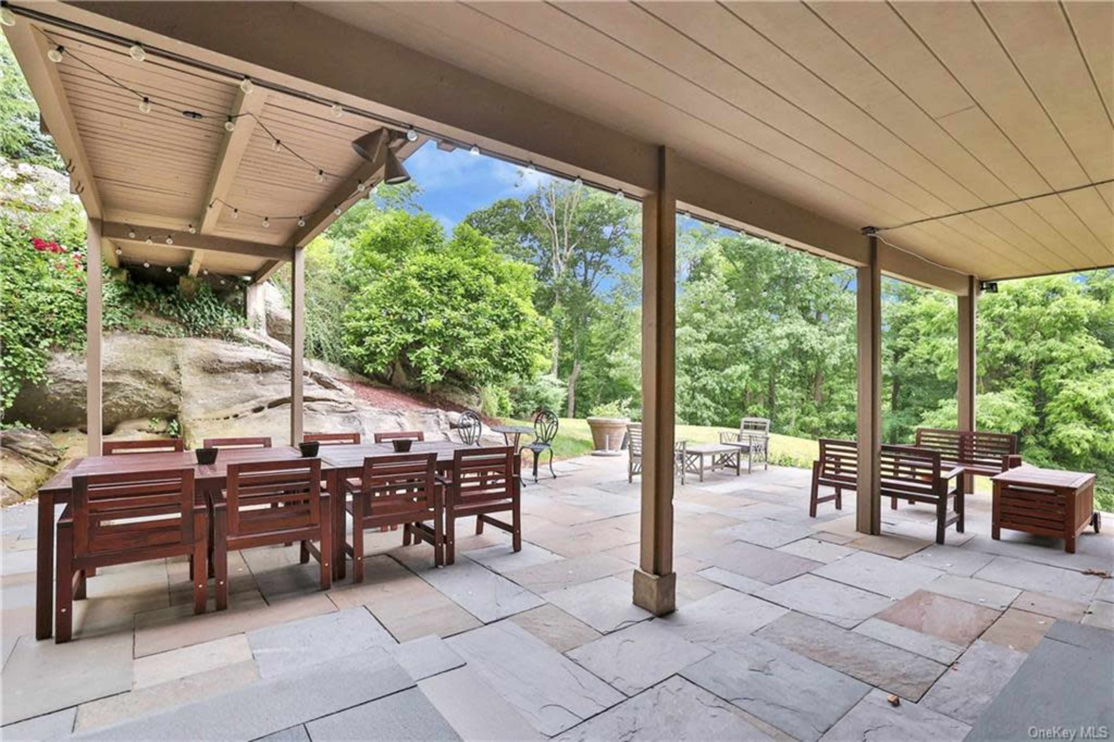 A covered outdoor patio with wooden dining tables and stone flooring, surrounded by greenery and rock formations.