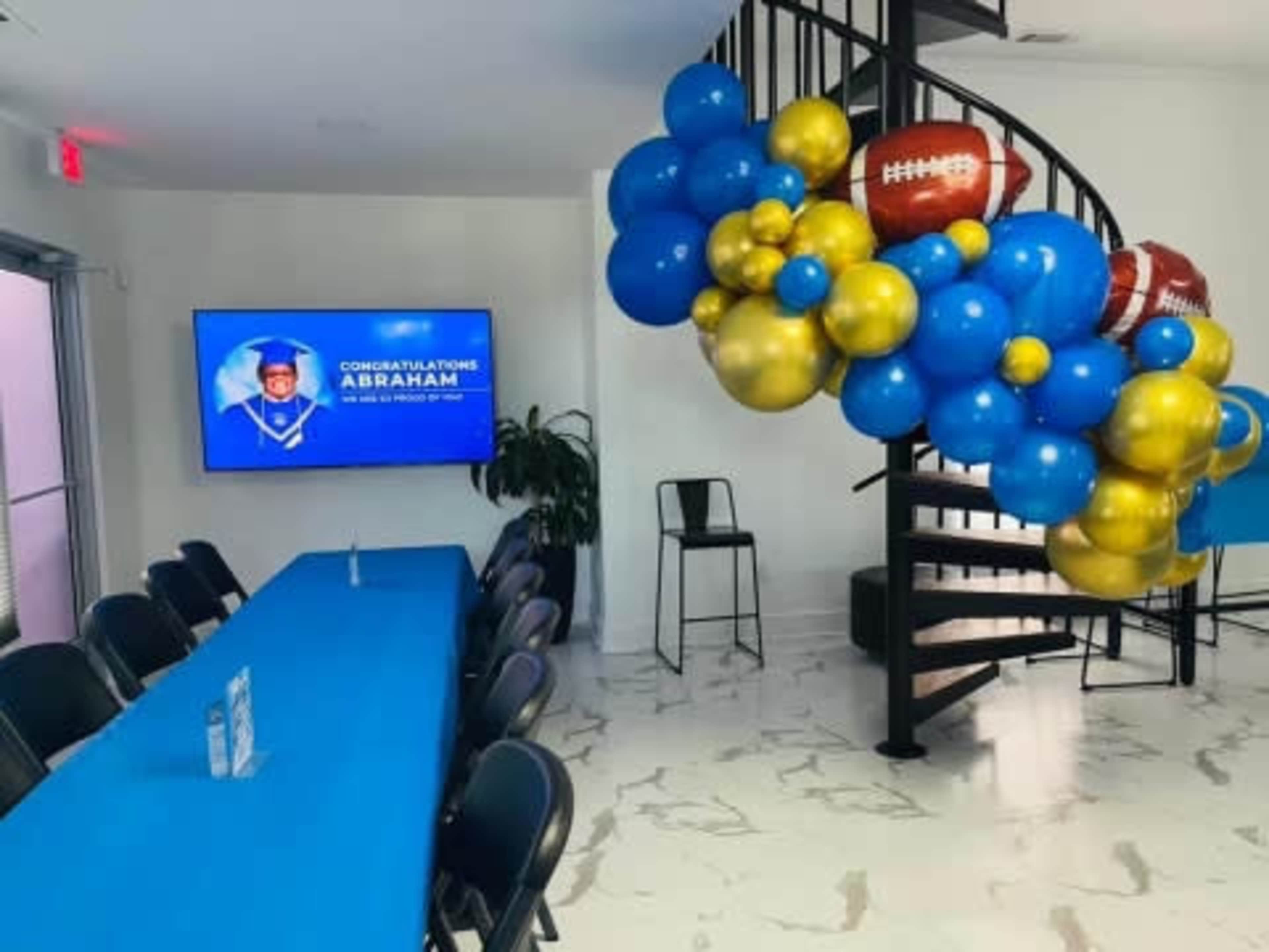 The image shows a decorated room with a blue and gold balloon arrangement featuring sports-themed decorations, a television screen displaying a congratulatory message for "Abraham," and a spiral staircase in the background.