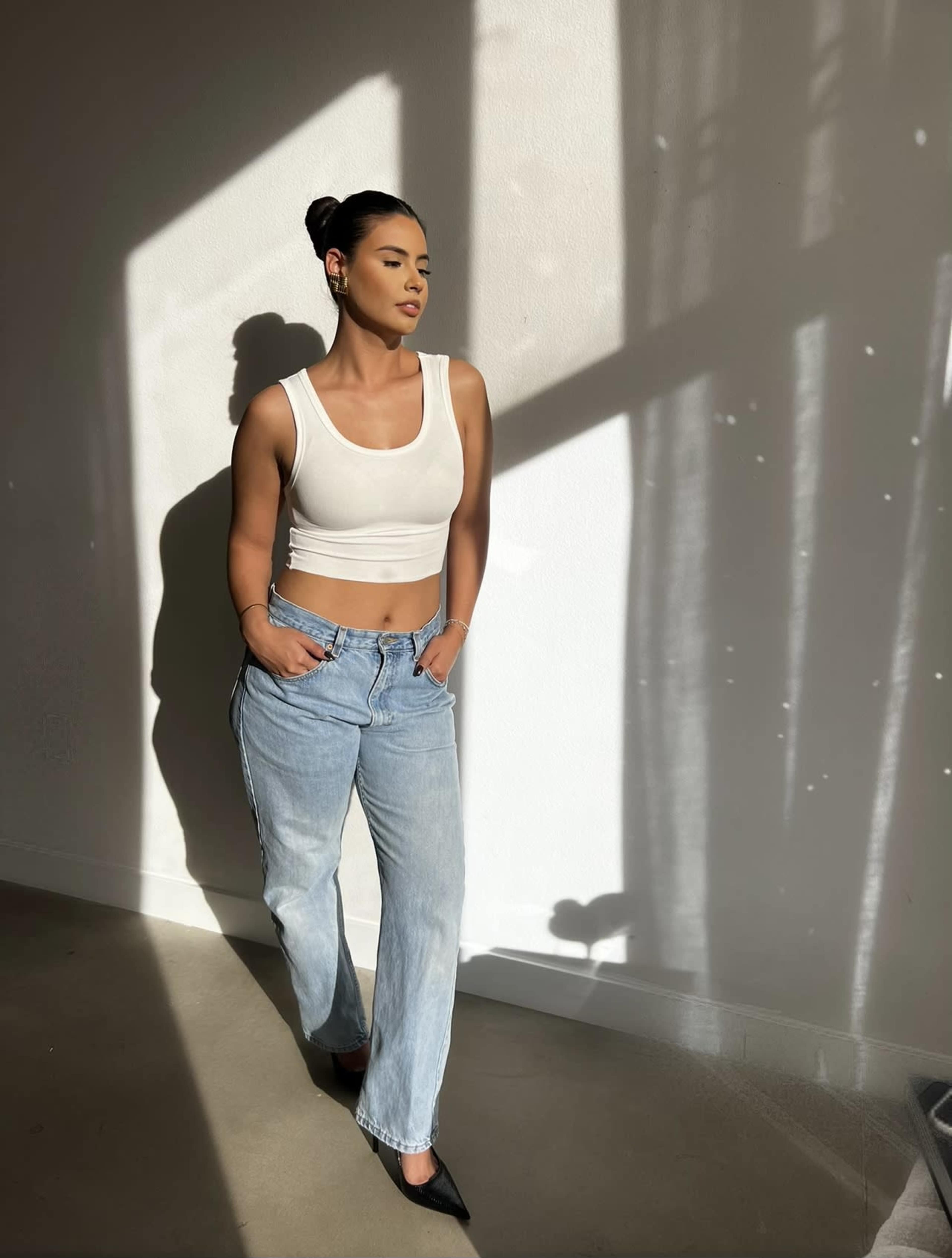 A woman wearing a white cropped tank top and light blue jeans stands against a wall with long shadows cast by natural light.