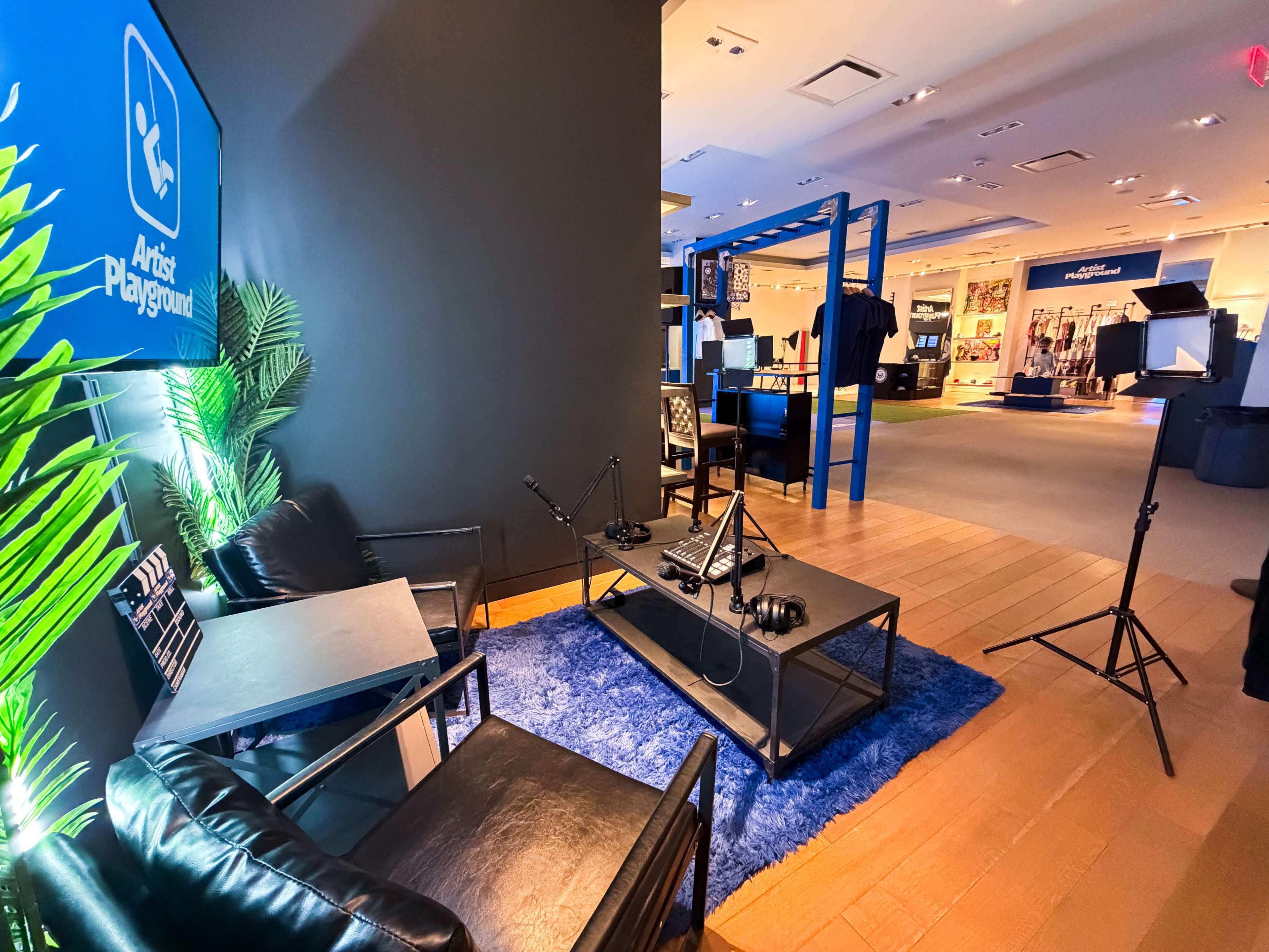 The image shows a stylish lounge area with black chairs and a glass coffee table, featuring two microphones and a headset on a table, illuminated by soft lighting in a retail space.