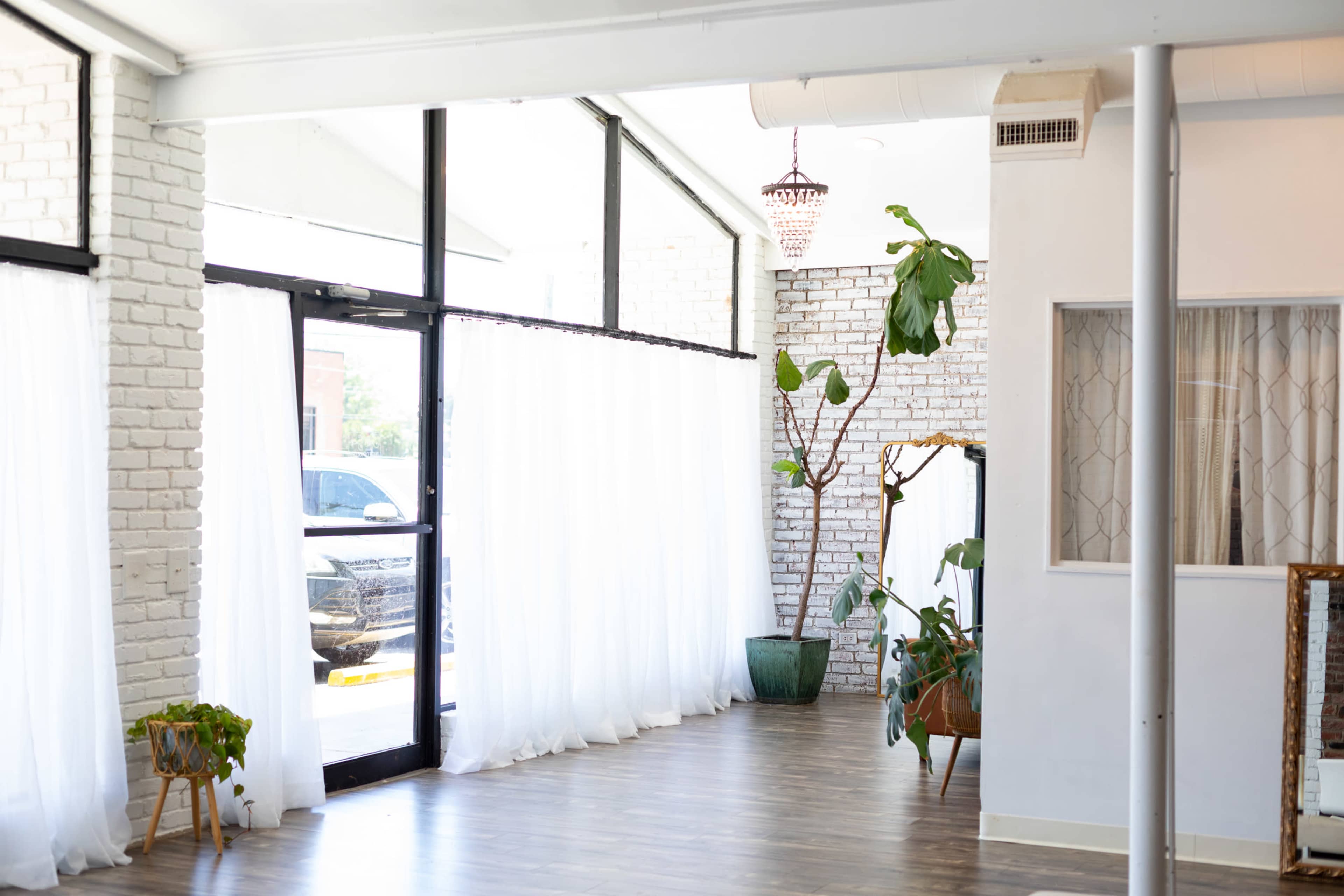 The image shows a spacious interior room with large windows, sheer curtains, potted plants, and wooden flooring.