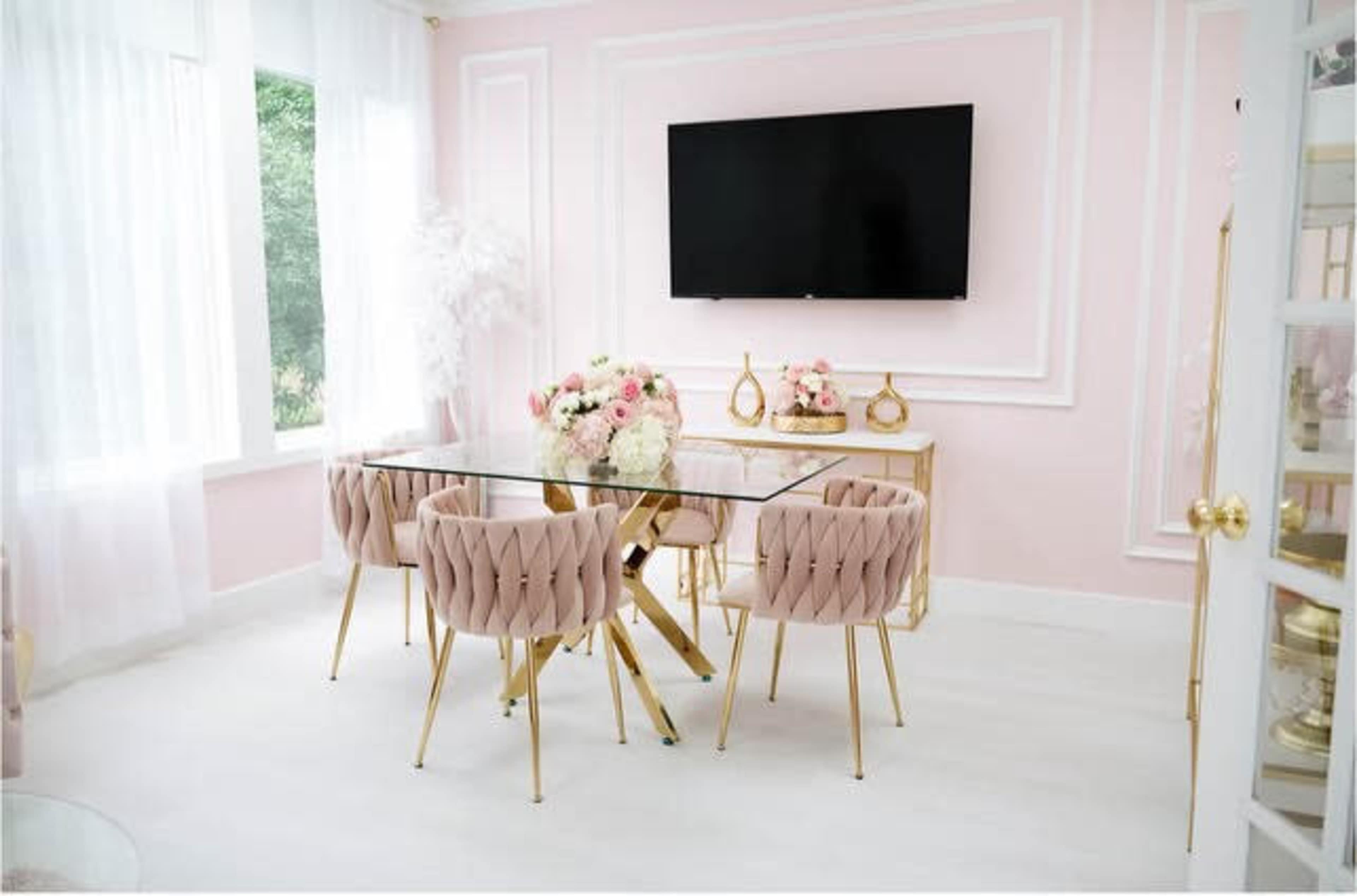 A modern dining area with a glass table surrounded by pink upholstered chairs and a large wall-mounted TV in a softly lit room with pink walls.