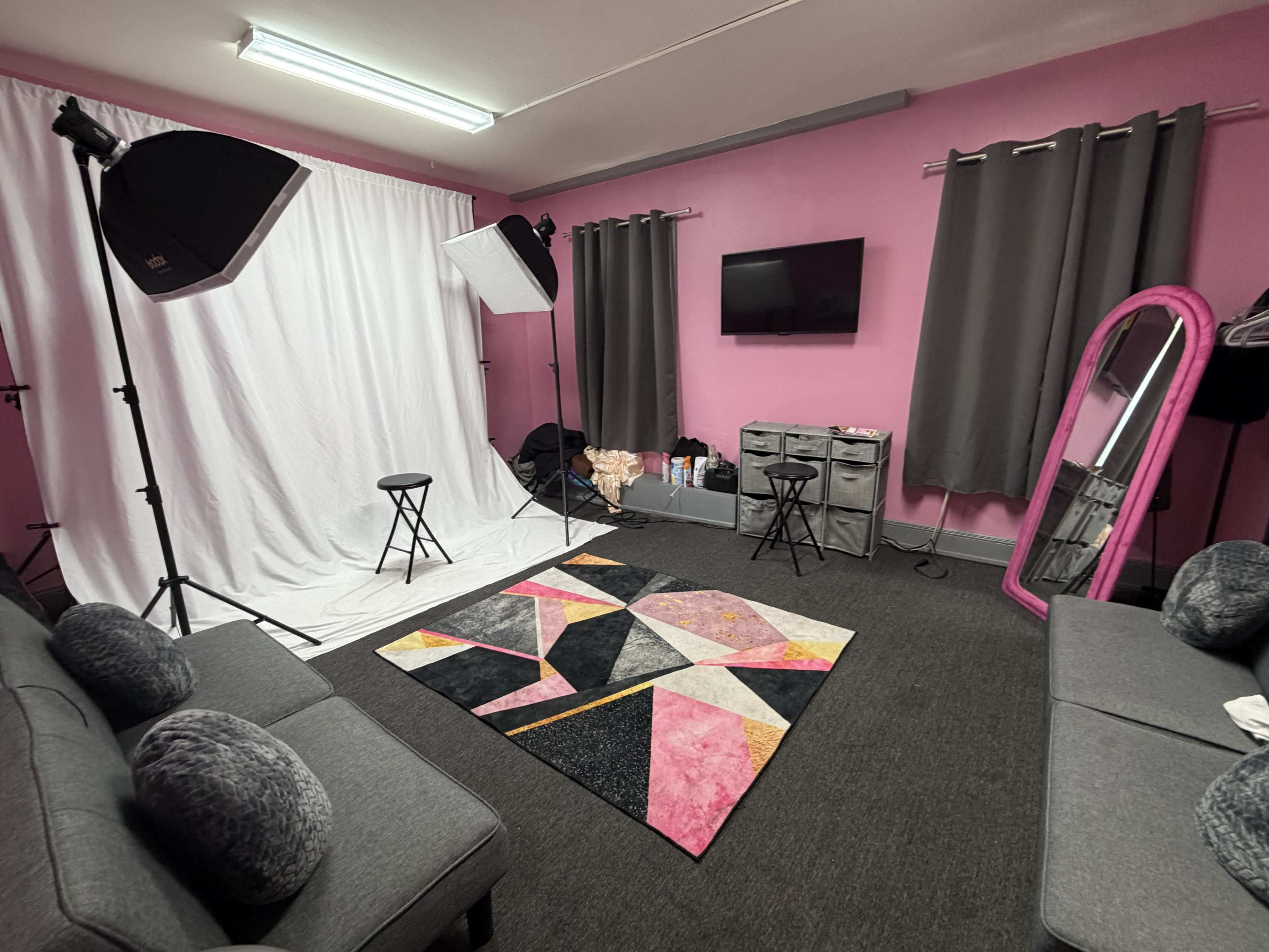 The image shows a small photography studio with pink walls, a white backdrop, softbox lights, a television, a rug with geometric patterns, and seating arranged for clients.