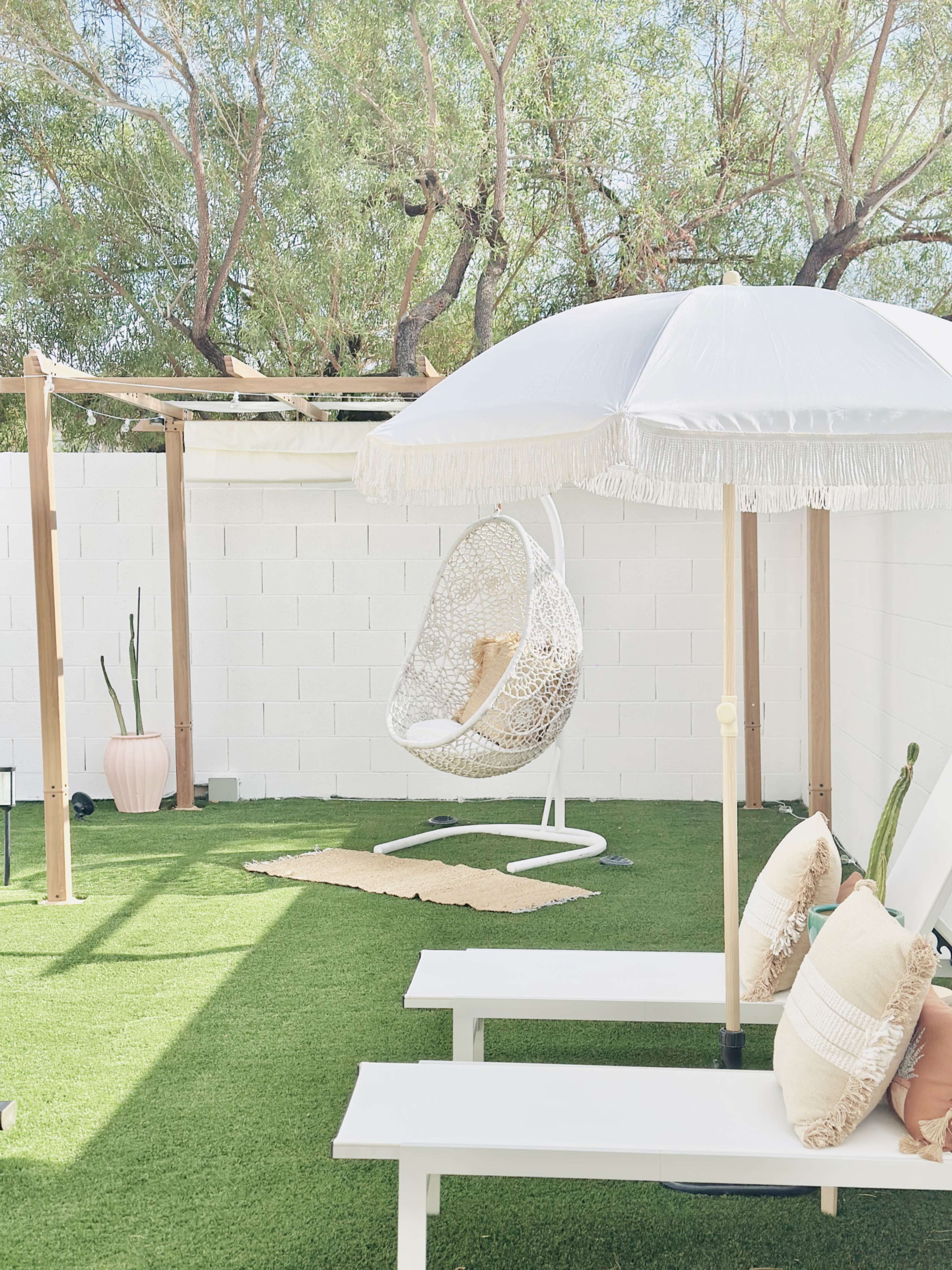 A well-lit outdoor space features a hanging chair, a shaded umbrella, and neatly arranged seating on artificial grass.