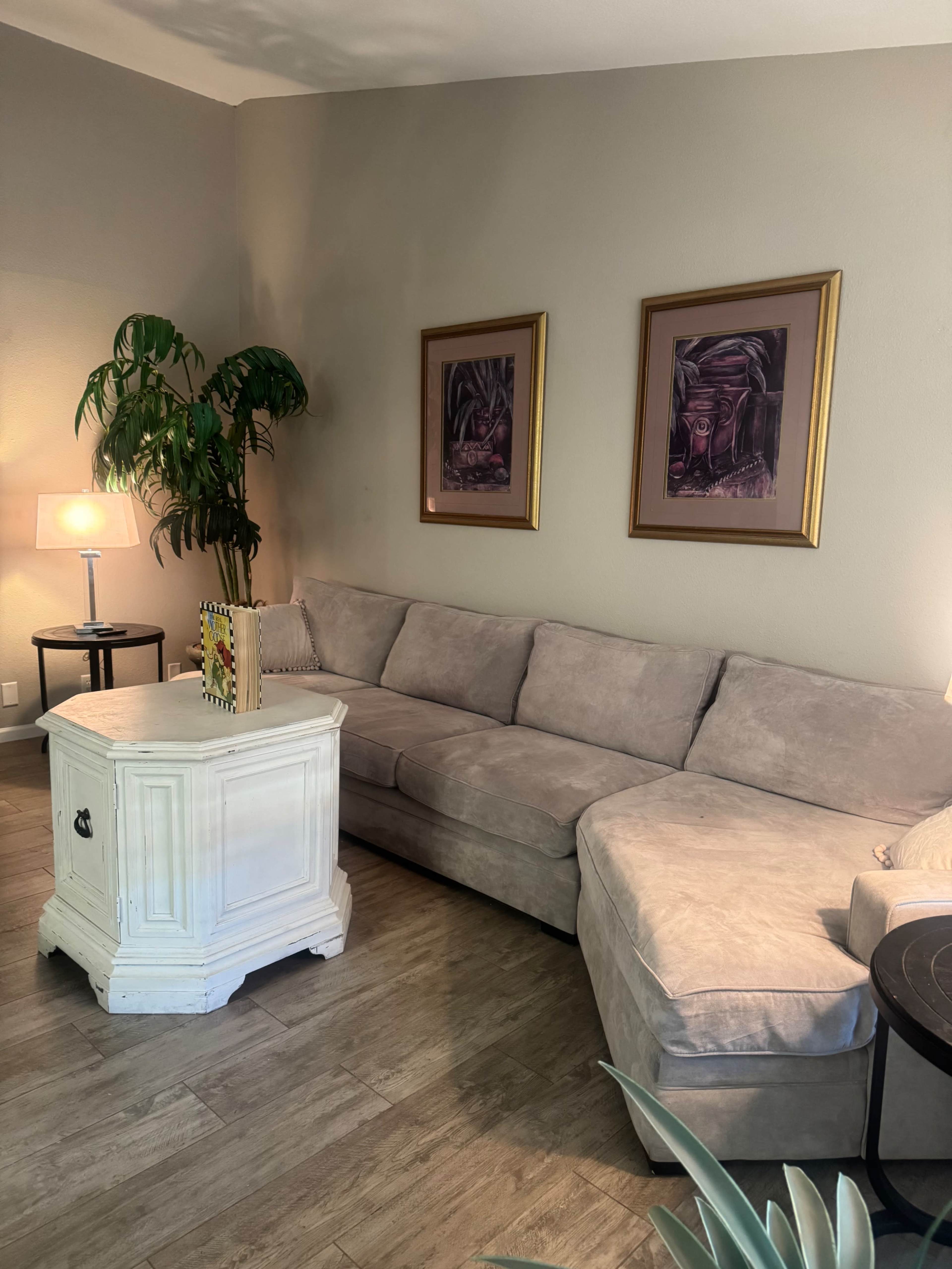 The image shows a cozy living room with a light-colored sectional couch, a small white table, a floor lamp, and two framed artworks on the wall, along with a potted plant in the corner.