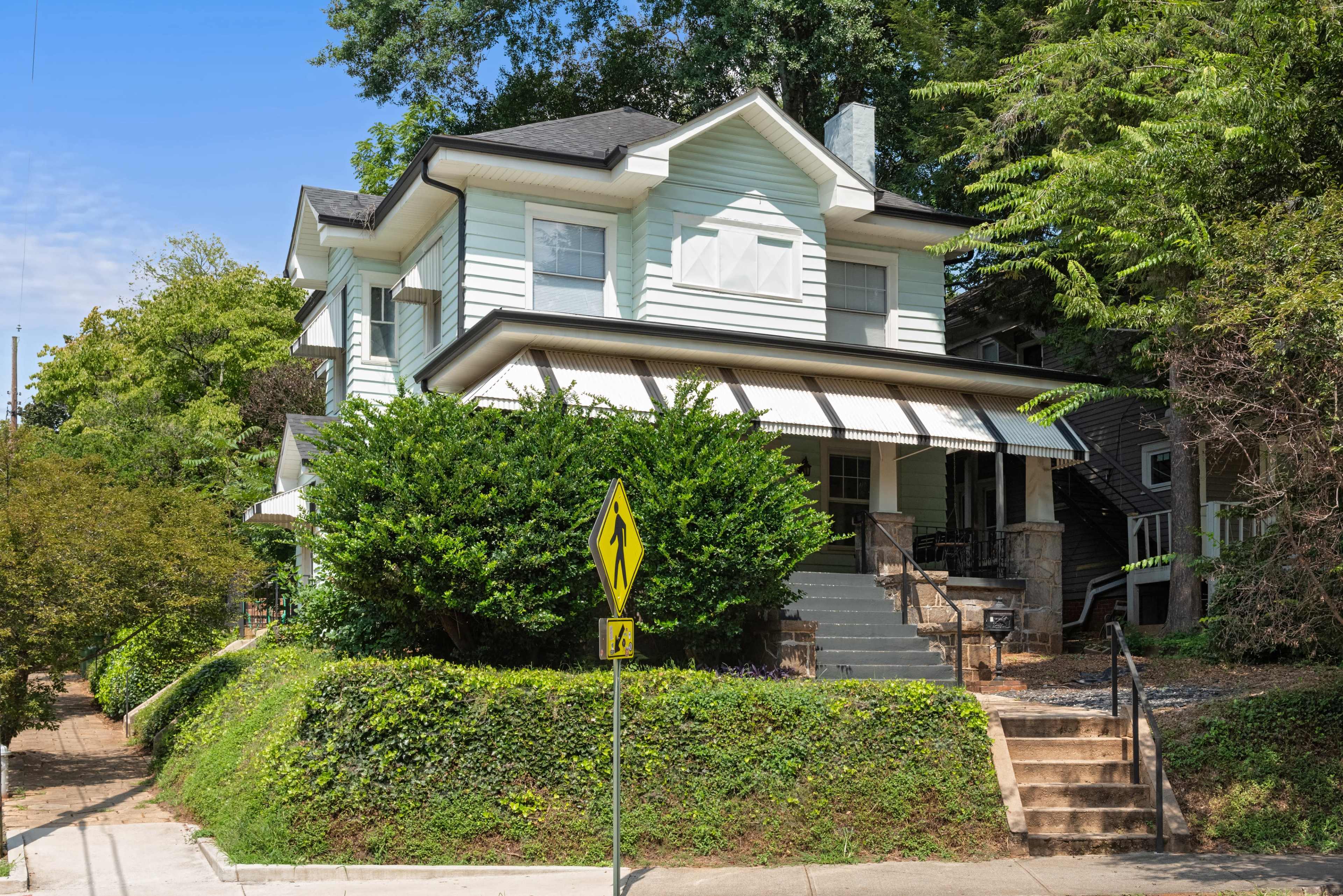 The image shows a light blue house with a front porch, surrounded by greenery and located on a sloped street with a pedestrian crossing sign nearby.