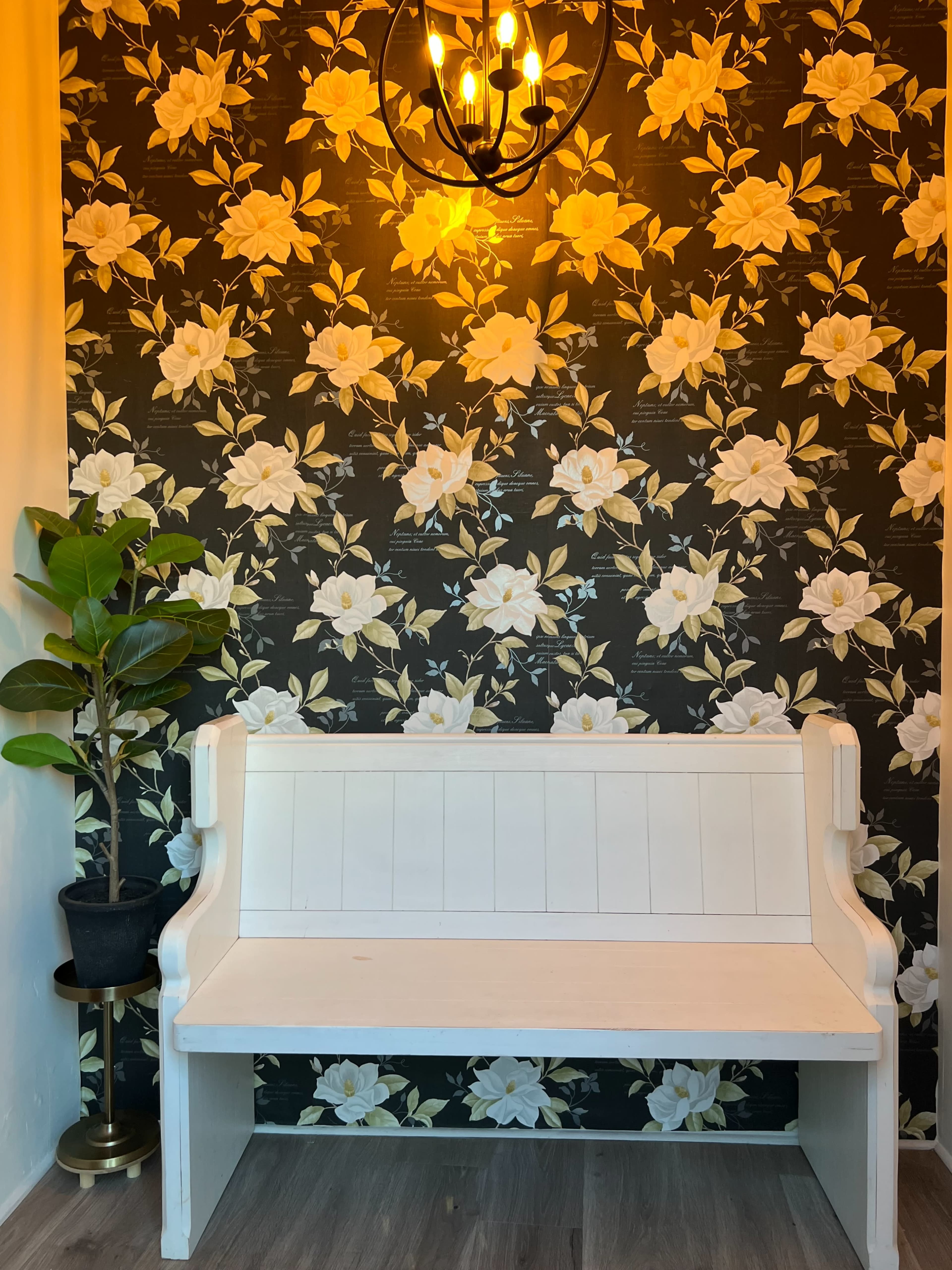 A white bench sits against a floral wallpaper backdrop, illuminated by a pendant light.