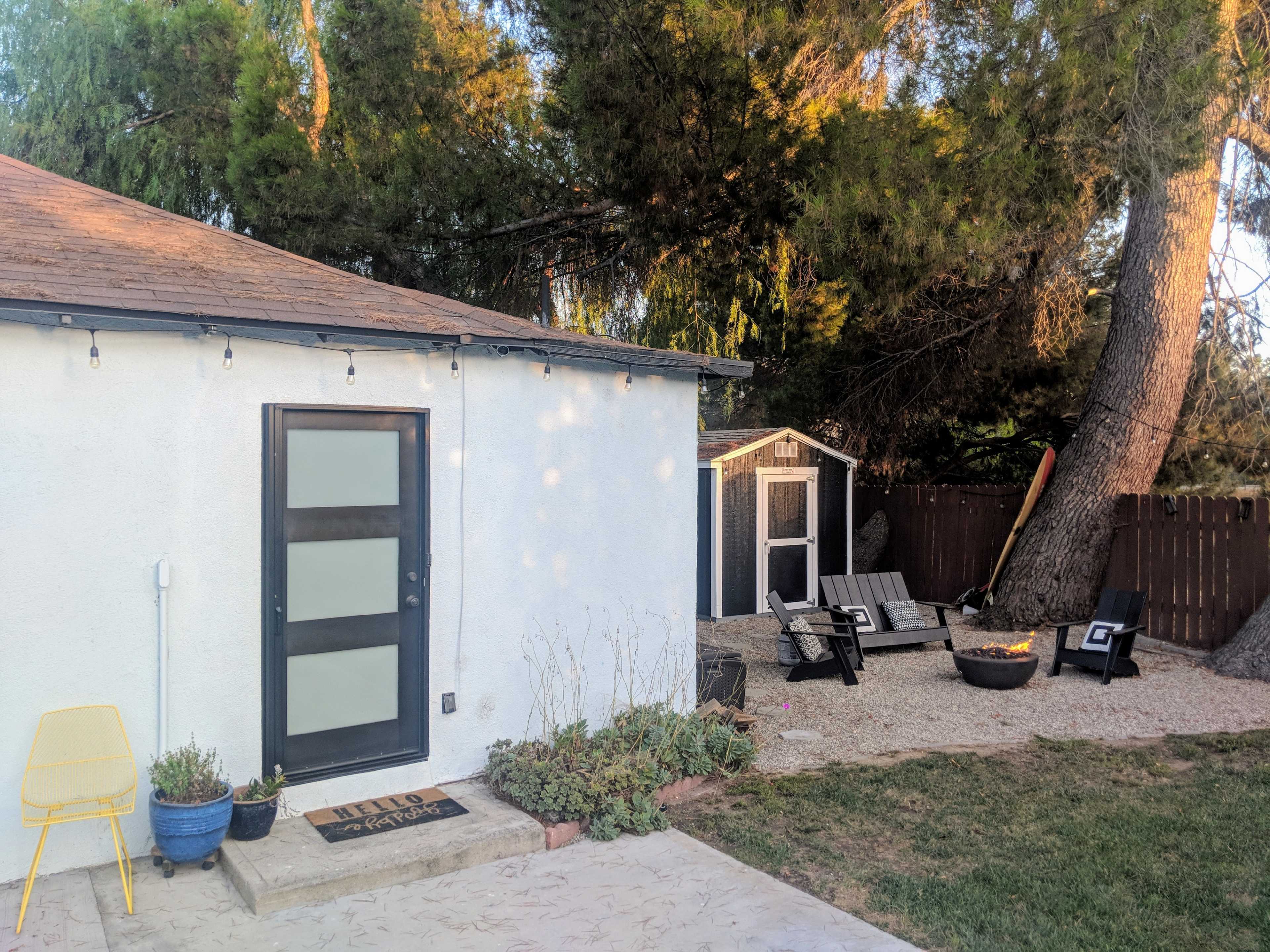 A small backyard features a white house, a shed, patio furniture, and a fire pit surrounded by gravel.