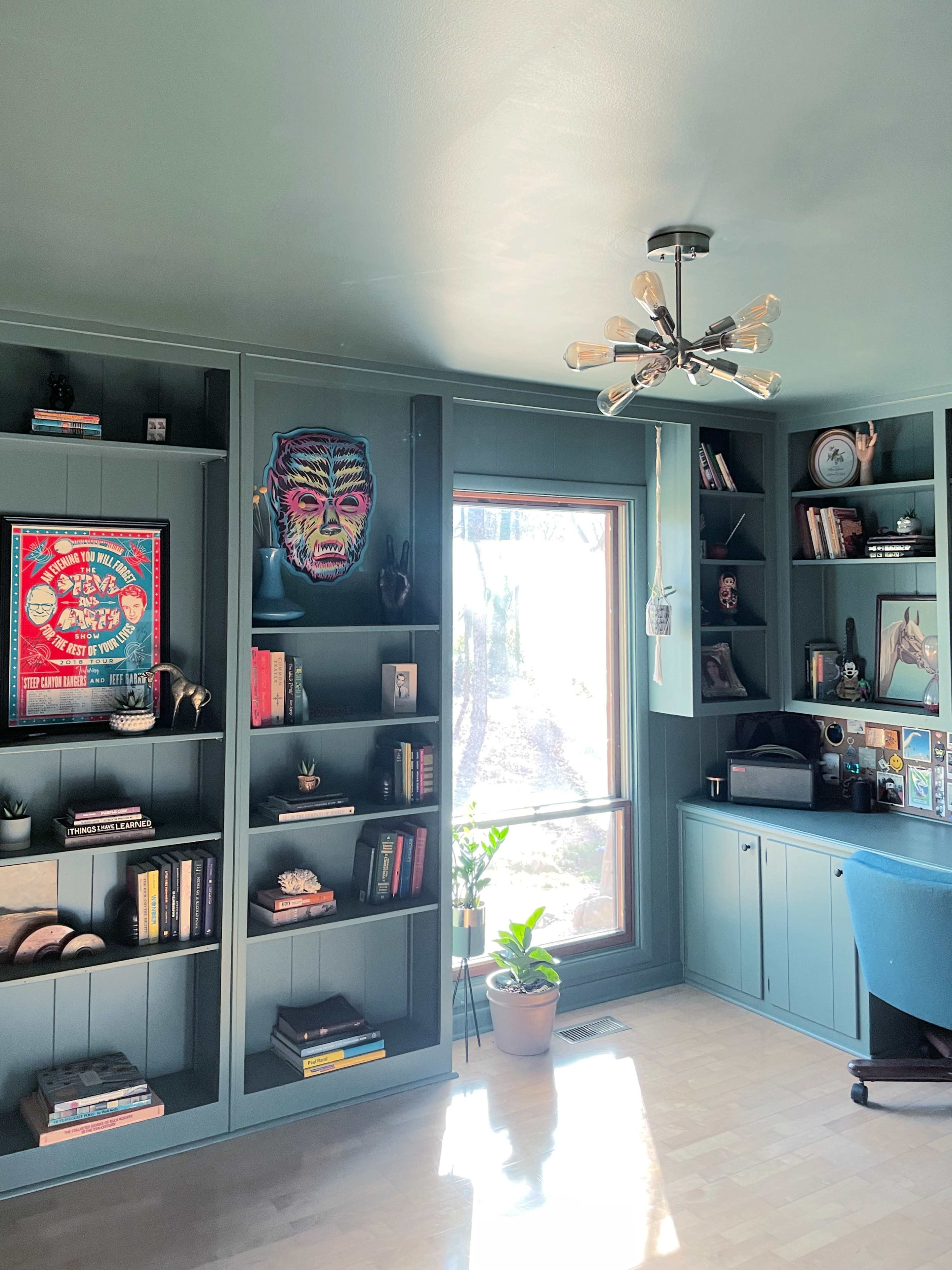 A well-lit room with dark teal walls and built-in shelves filled with books and decorative items, featuring a large window with a view of greenery outside.