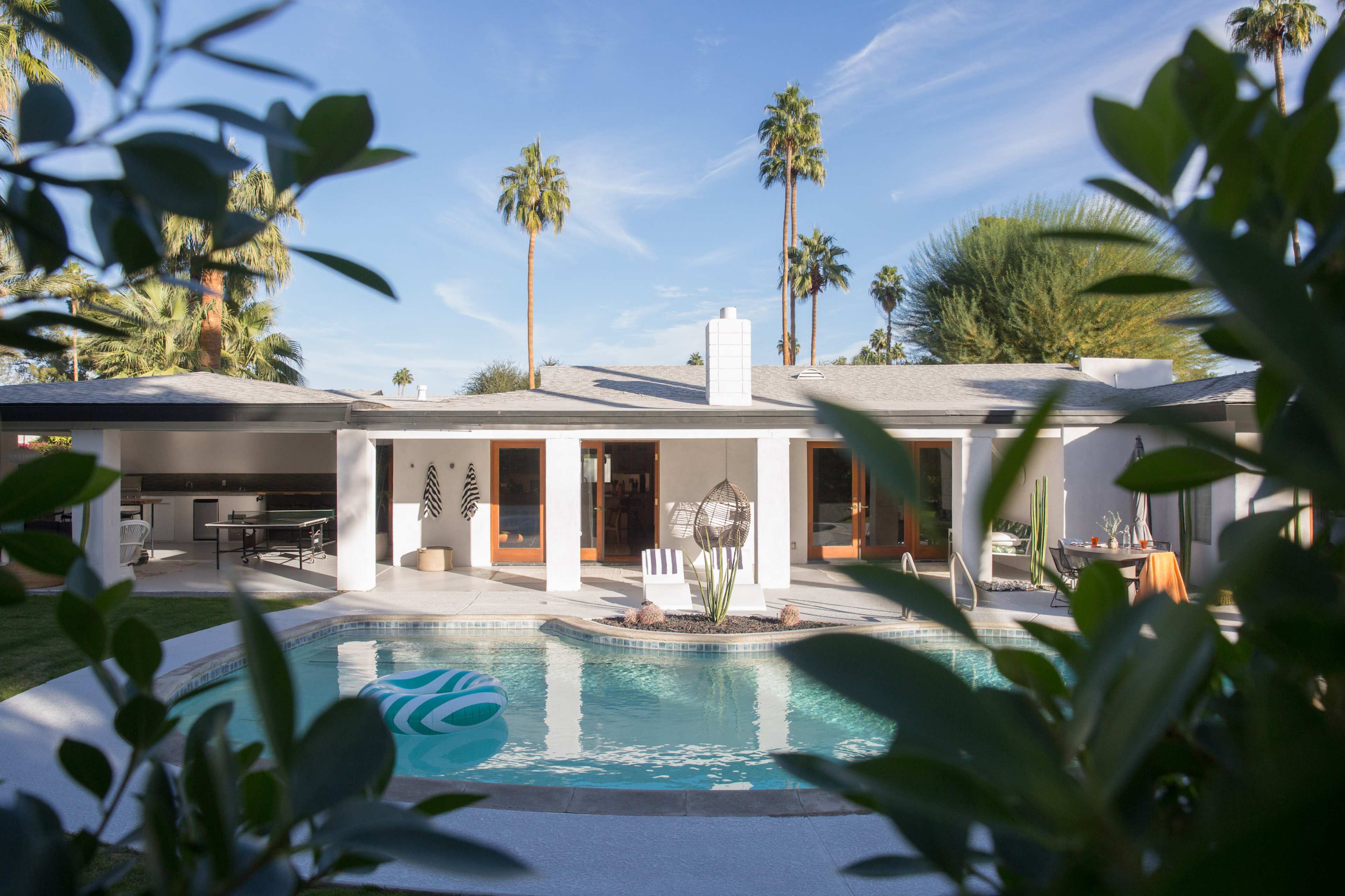A modern house with large windows overlooks a pool surrounded by palm trees.