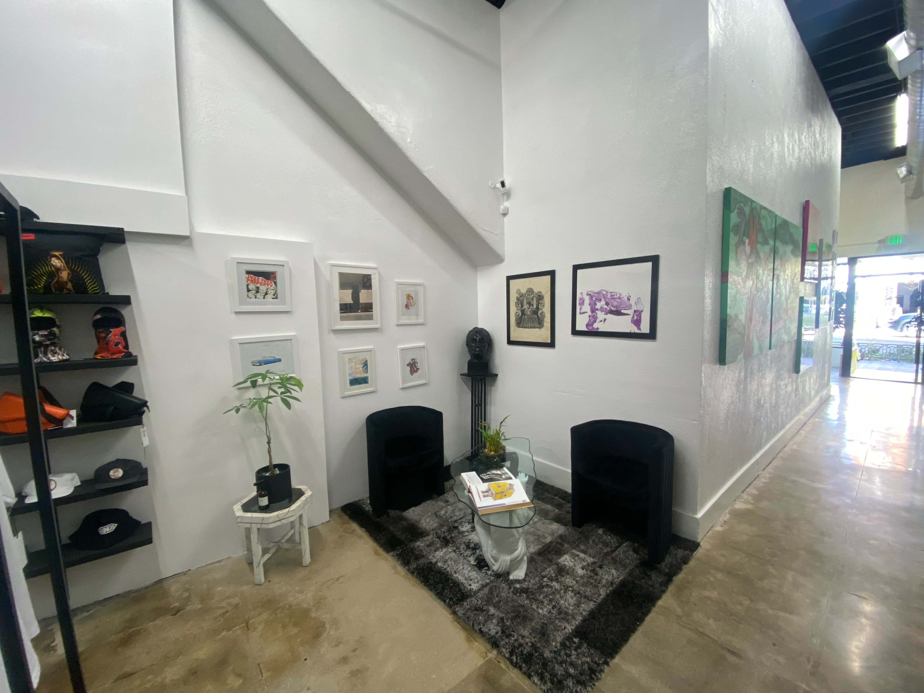 The image shows a corner of a gallery or studio featuring two black chairs, a small glass table, a plant, framed artwork on the wall, and shelves displaying hats.