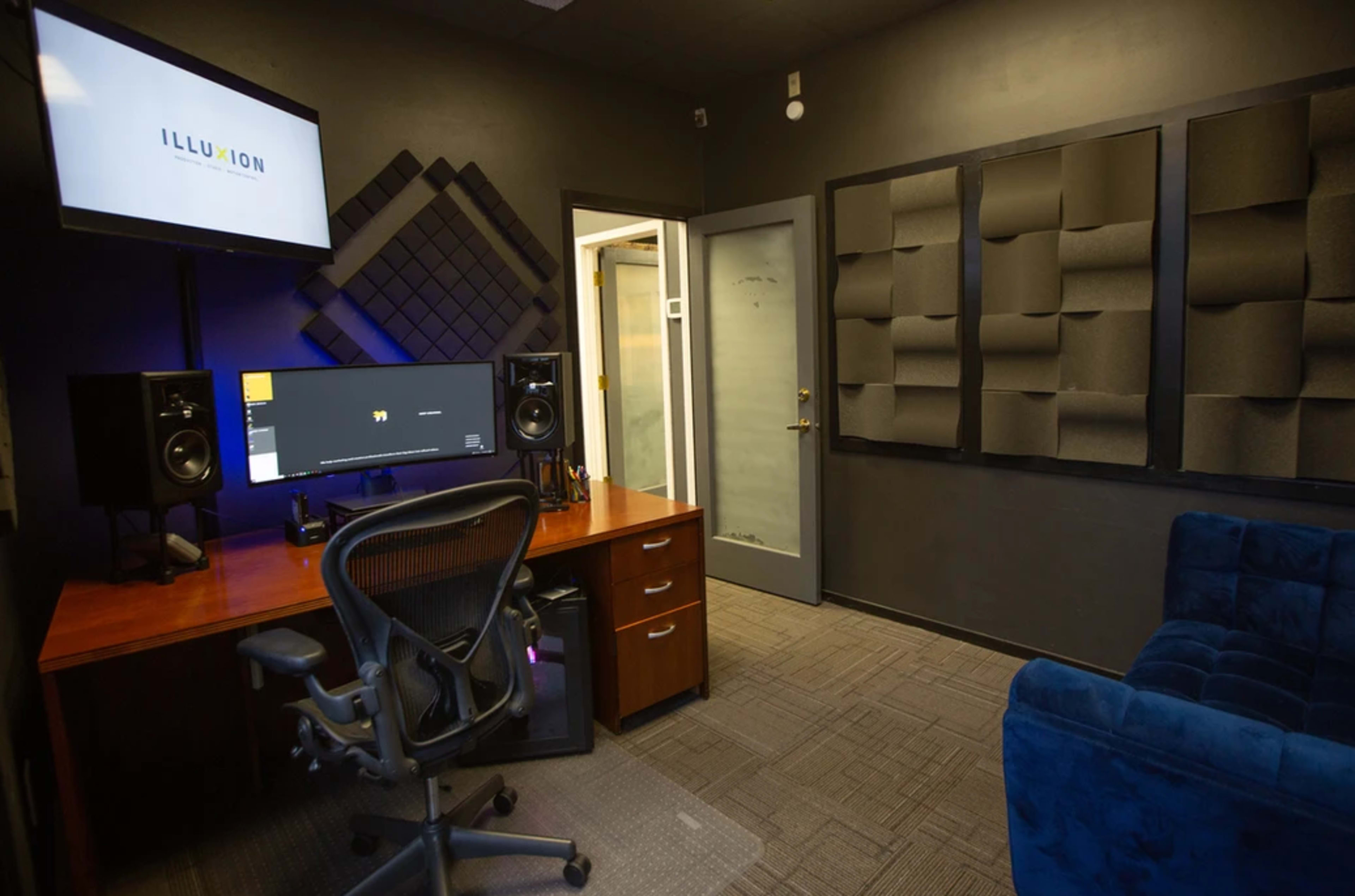 The image shows a small recording studio featuring a desk with a computer monitor, speakers on either side, soundproof wall panels, and a blue couch.