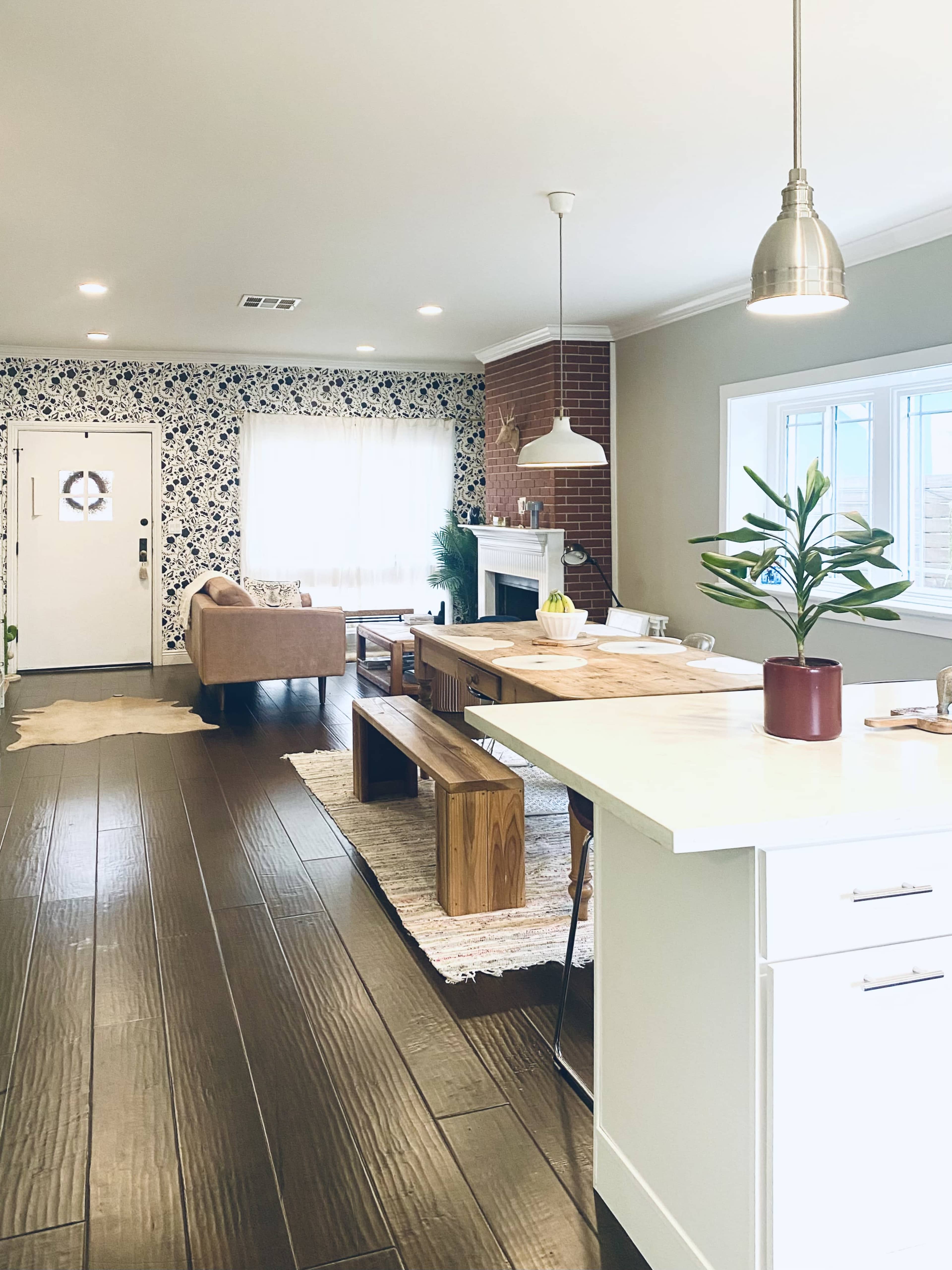 The image shows a modern, open-concept living space featuring a dining area with a wooden table, a beige couch, and a fireplace against a floral wallpaper backdrop.