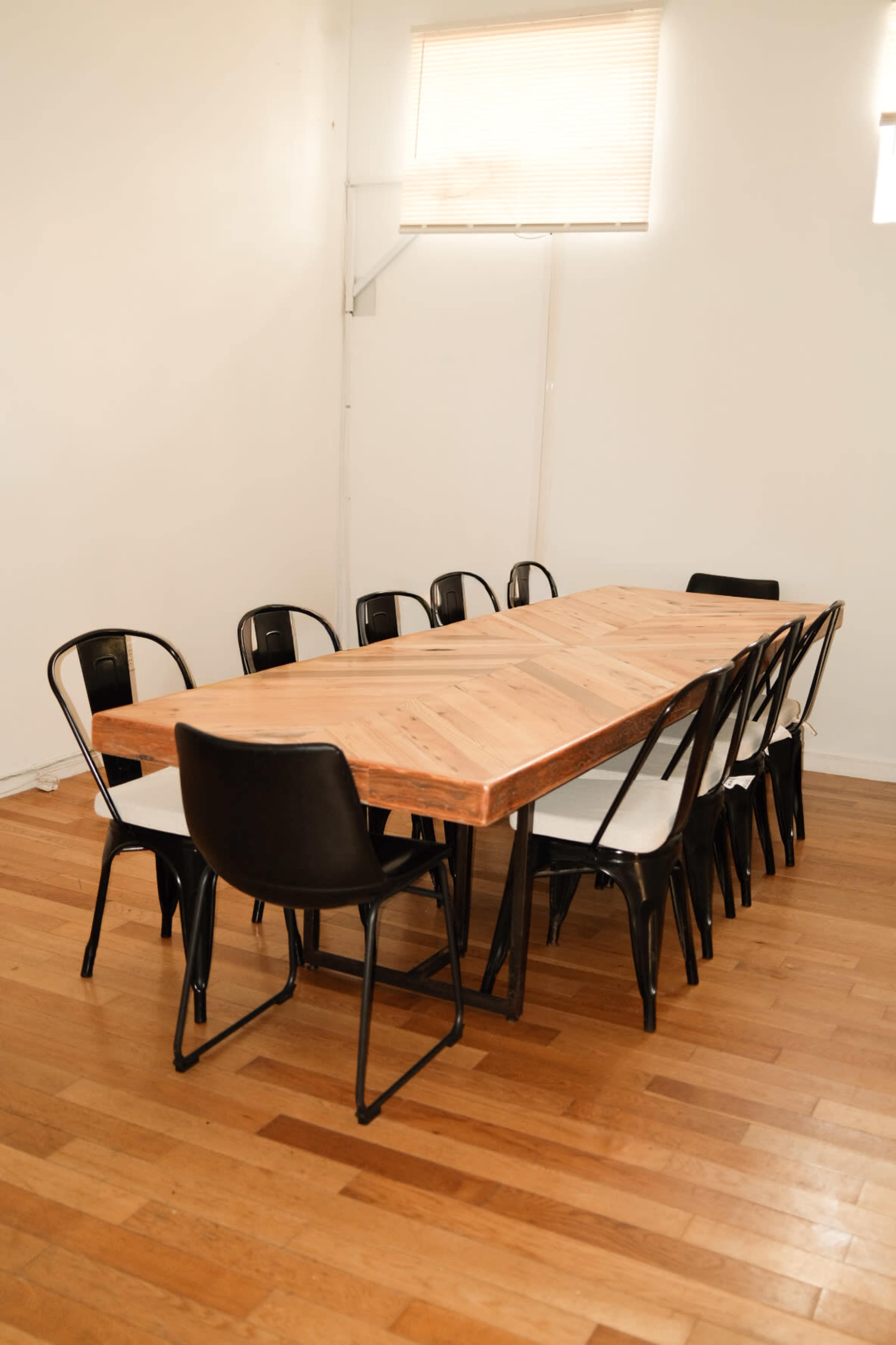 A long wooden table surrounded by black metal chairs is situated in a bright, minimalist room with white walls and hardwood flooring.