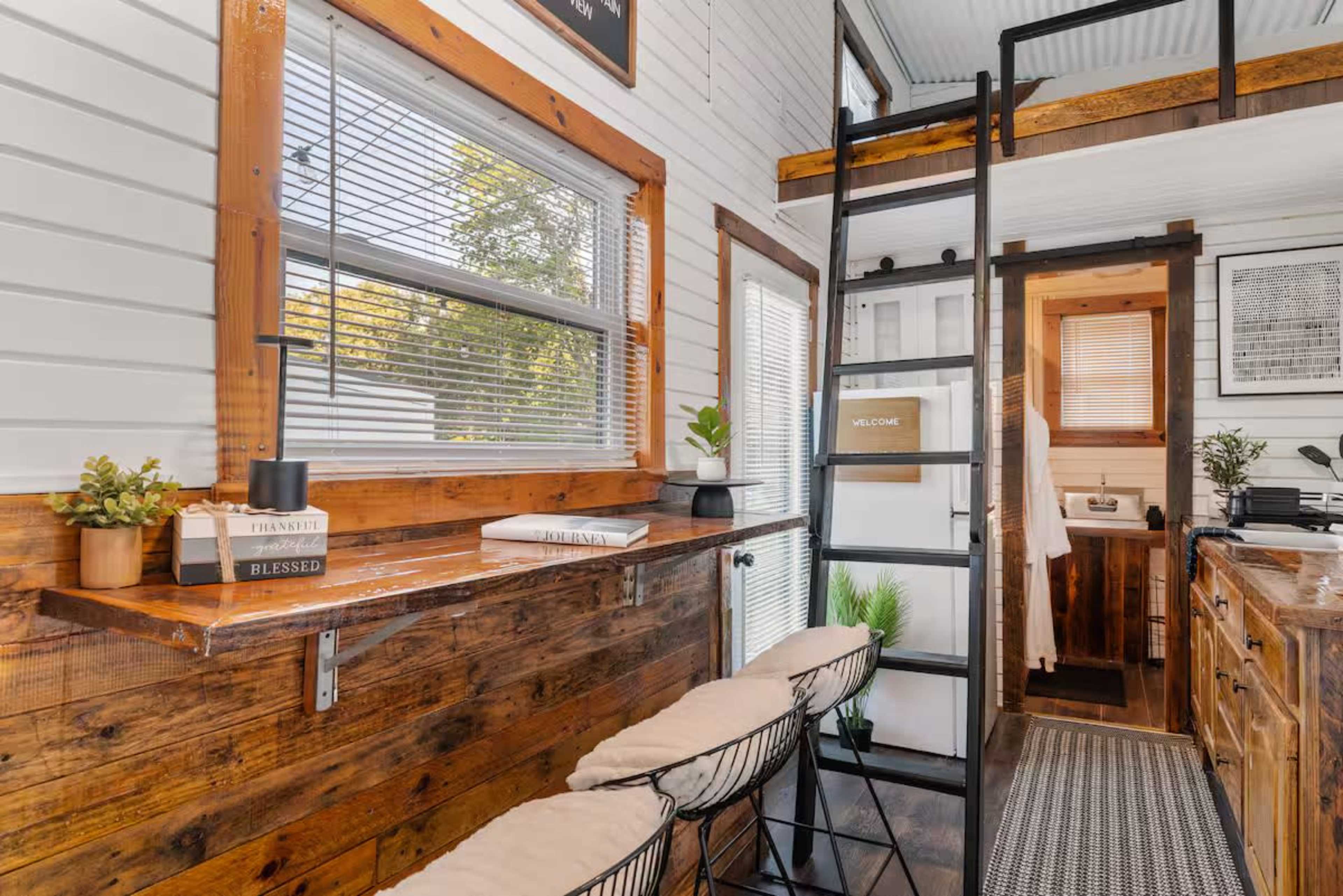 The image shows the interior of a tiny house featuring wooden accents, a lofted sleeping area accessible by a ladder, and a cozy seating area with bar stools.