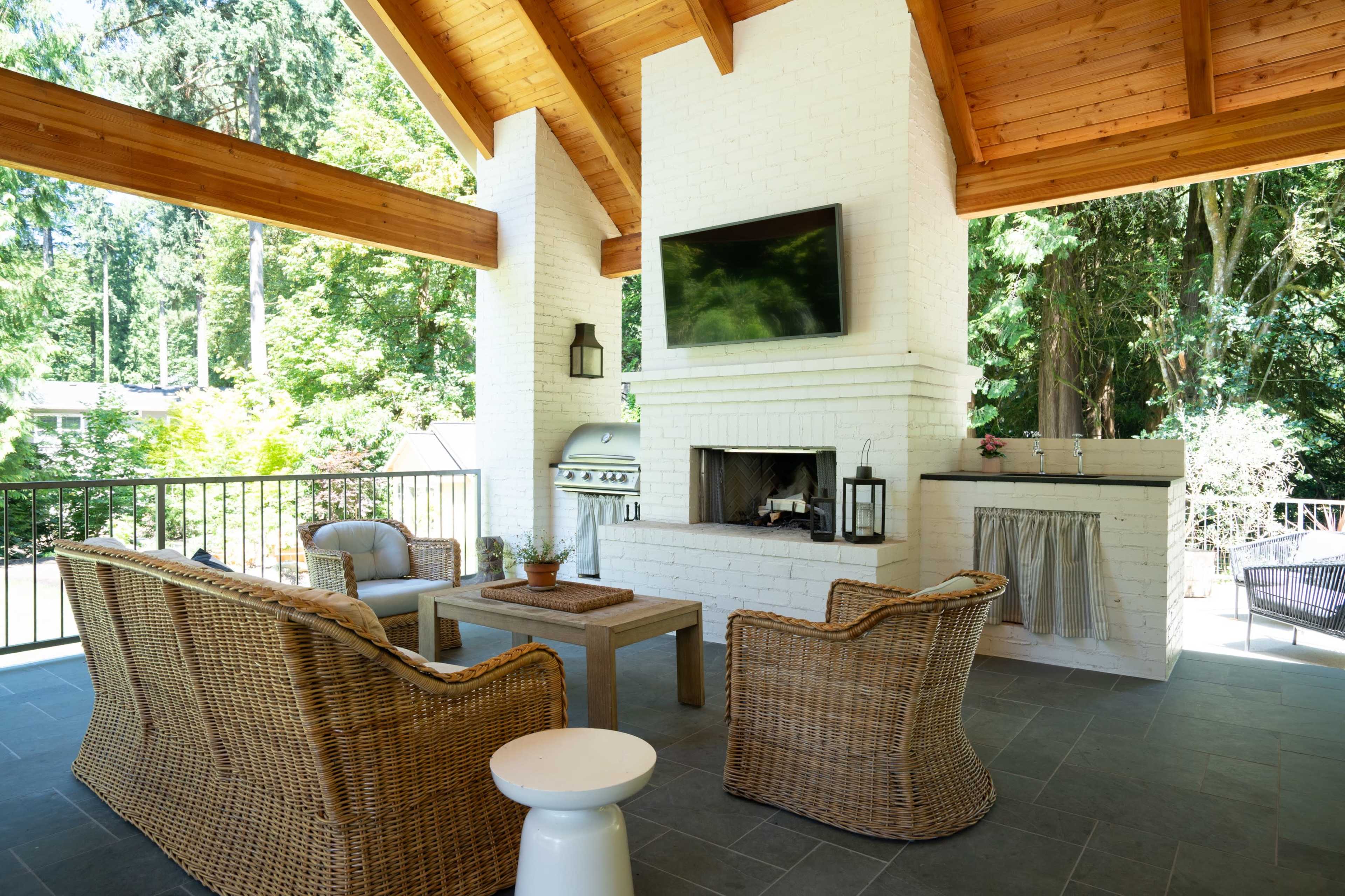 The image shows a spacious outdoor patio with a brick fireplace, a grill, seating area with wicker chairs, and a television mounted on the wall, all surrounded by greenery.