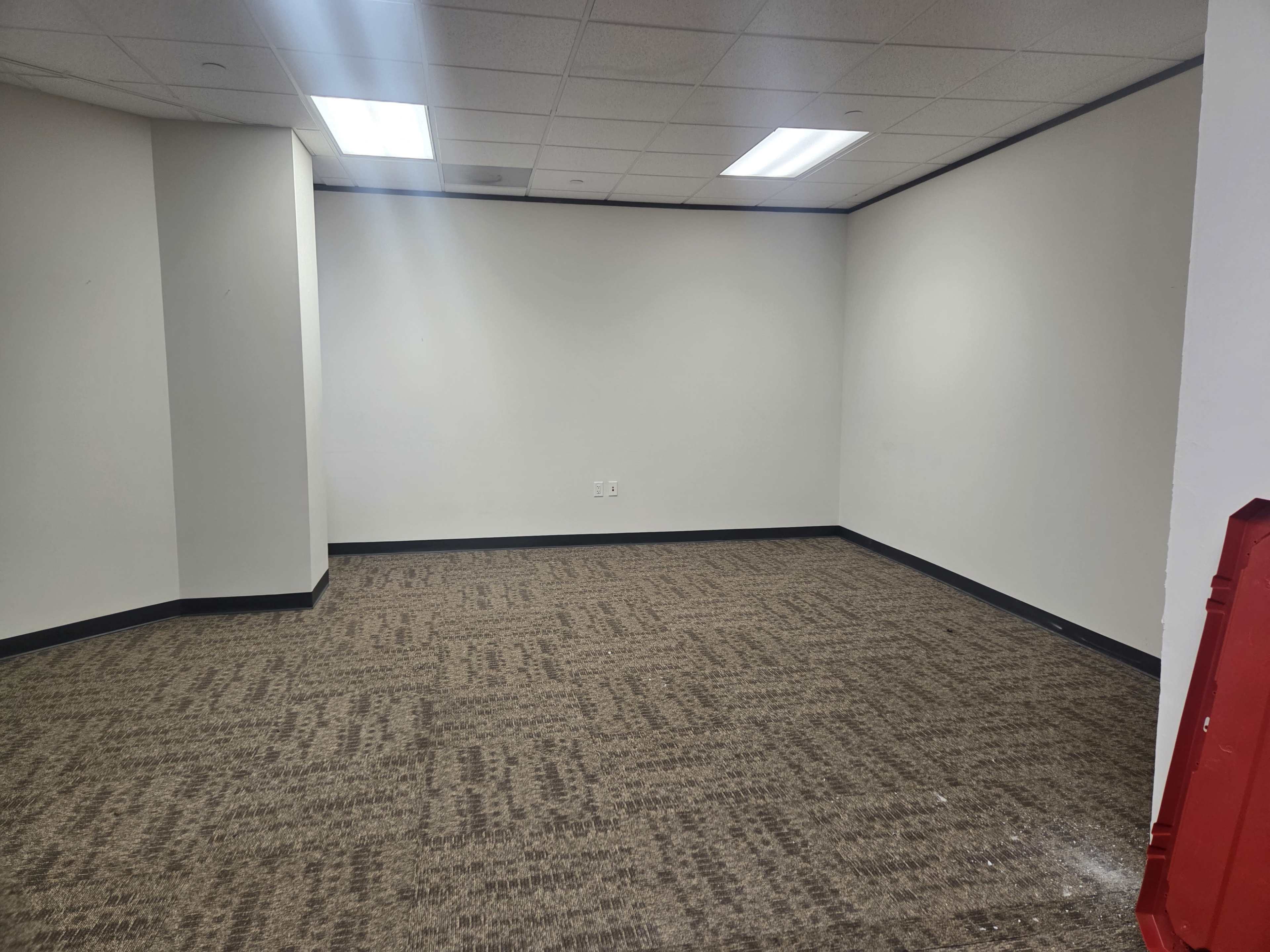 The image shows a vacant room with a carpeted floor, white walls, and fluorescent lighting.